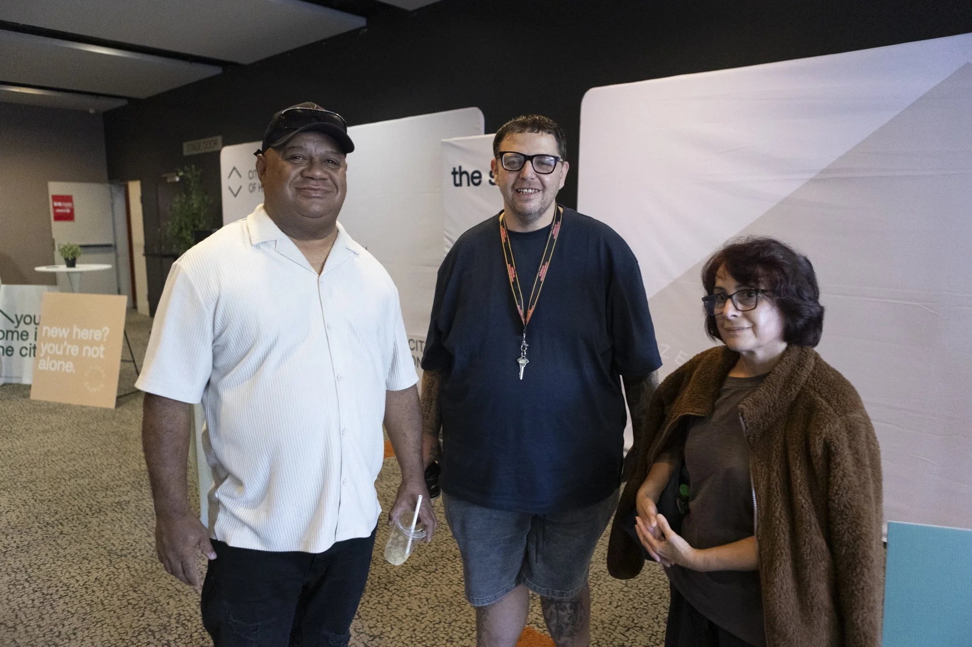 Three people standing indoors, smiling at the camera. The person on the left is a man wearing a white shirt, black cap, and holding a drink. The person in the middle is a man with glasses, wearing a black T-shirt and shorts. The person on the right is a woman with glasses, wearing a brown coat and holding a water bottle.