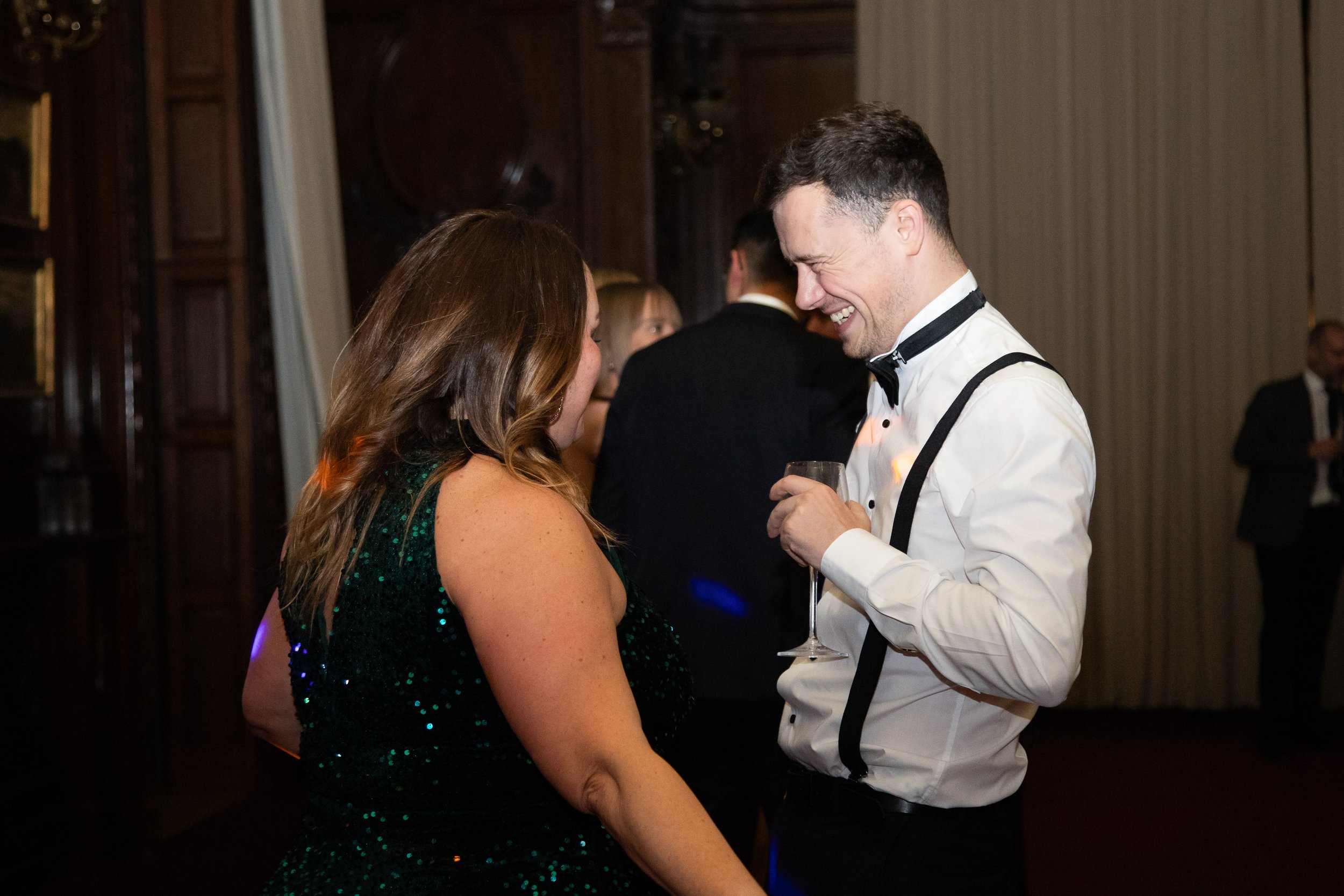 A man and woman are dancing and smiling at a formal event. The man is wearing a tuxedo with suspenders and bow tie, holding a glass of wine. The woman has long brown hair and is wearing a dark green, sequined dress. They are close together, enjoying 