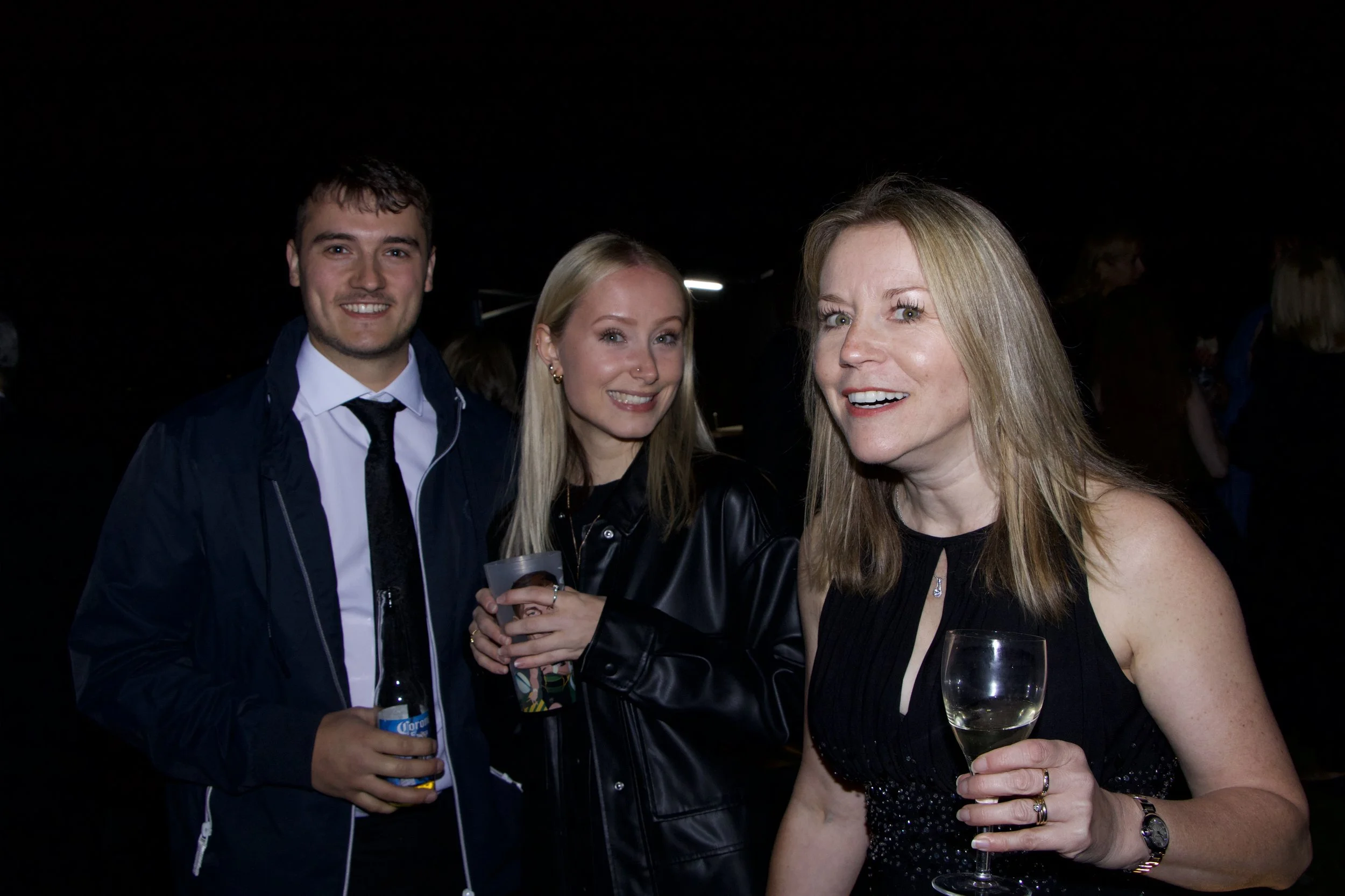 Three people at a social event, with two women and one man, holding drinks and smiling at the camera. The woman on the right has long blonde hair and is holding a glass of wine. The woman in the middle has blonde hair and is holding a drink. The man 
