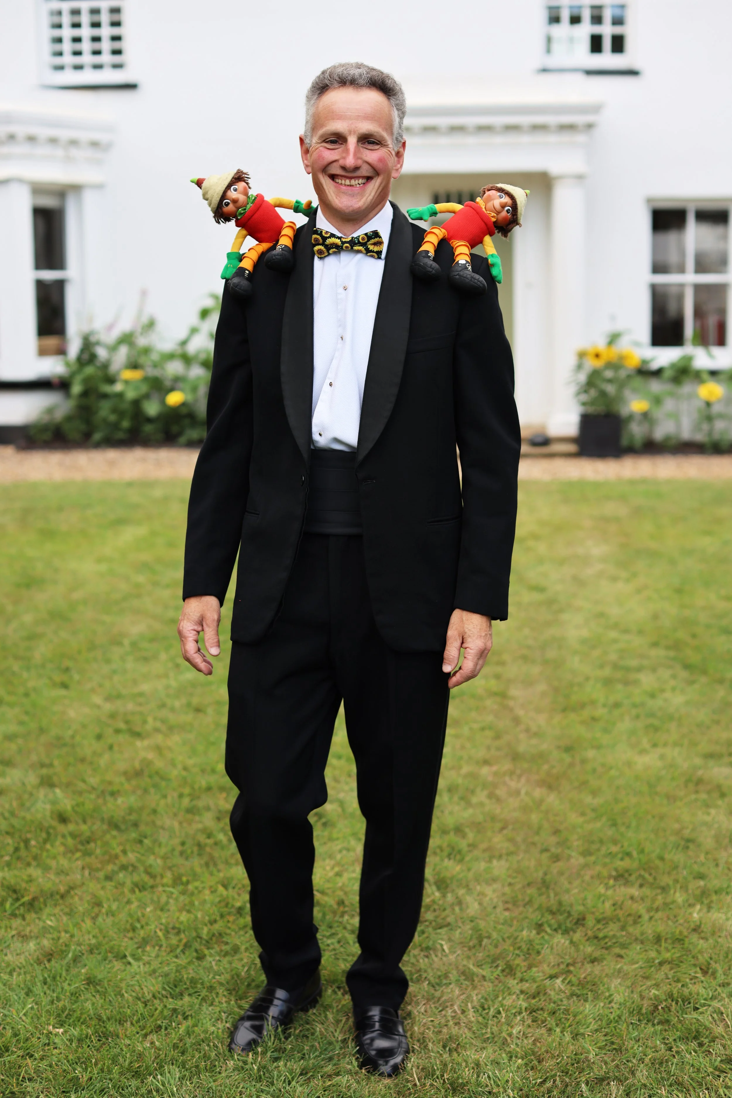 A man in a tuxedo with a bow tie, smiling, standing outside on grass with a house in the background. He has two puppets with elf-like hats perched on his shoulders.