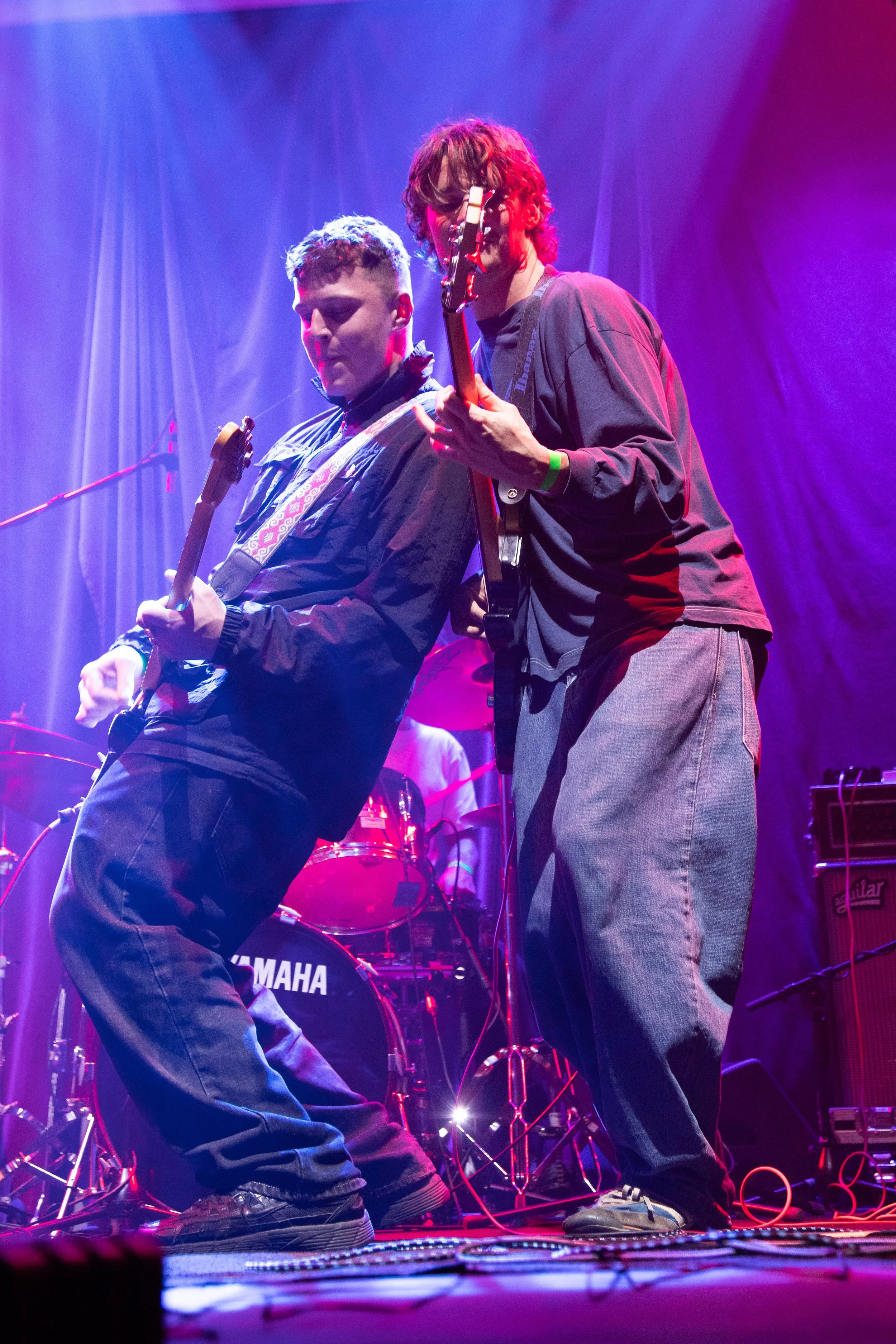 Two musicians performing on stage with colorful lighting, one playing an electric guitar and the other possibly singing into a microphone, with a drum set and musical equipment in the background.