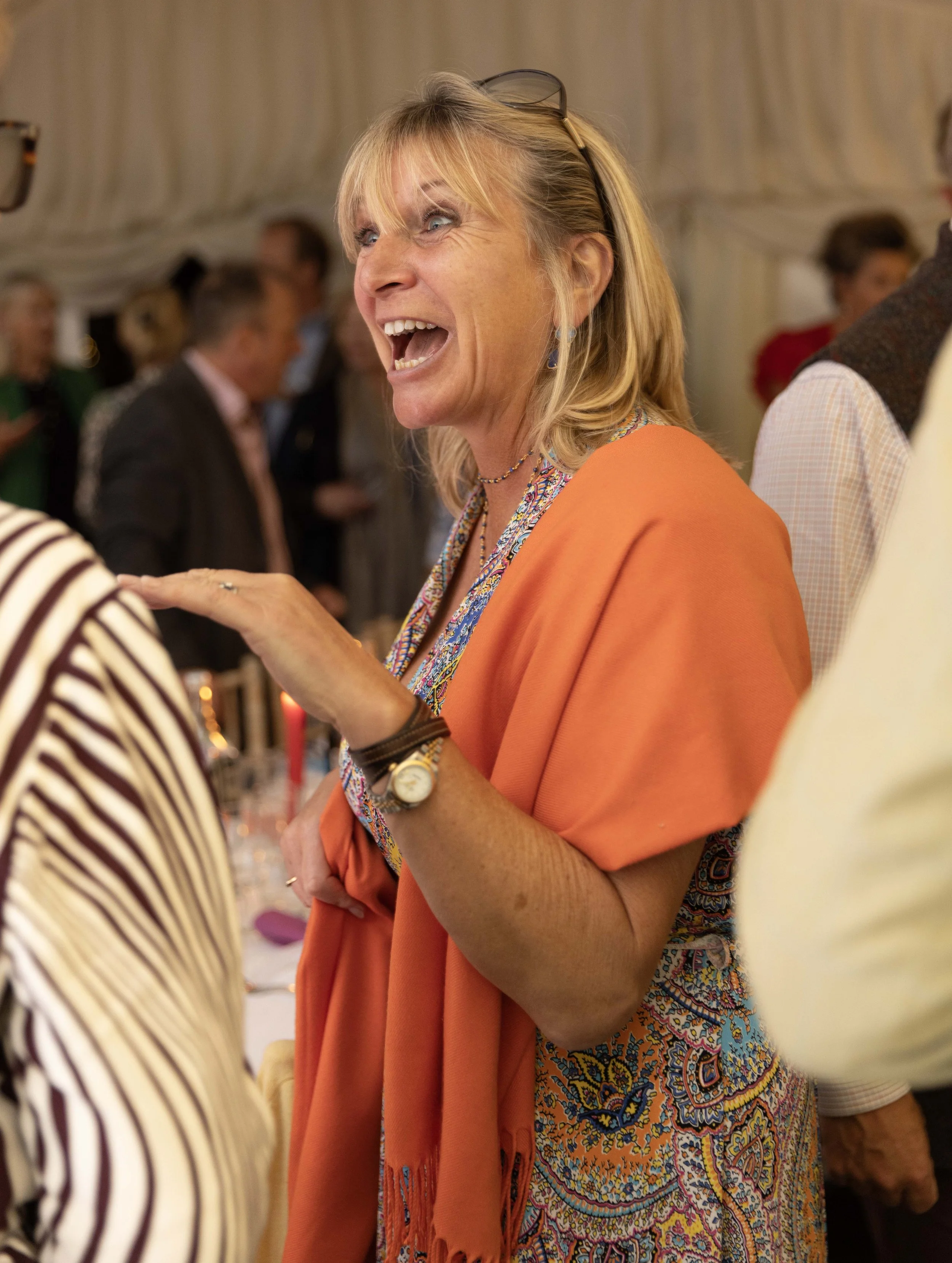 A woman with blonde hair, wearing glasses on her head and a colorful dress, is laughing and engaging in conversation at a social gathering.