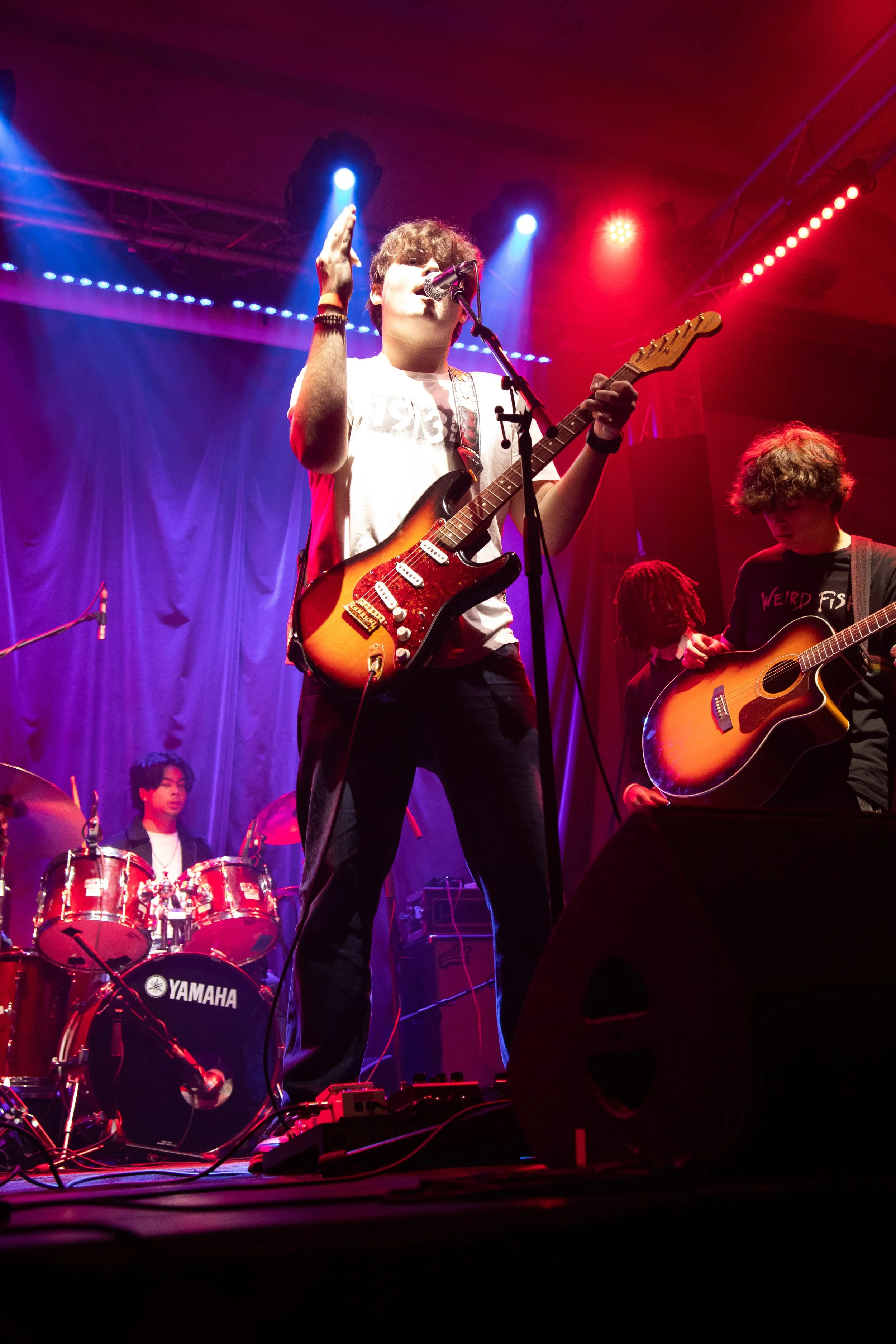 Musician performing on stage with electric guitar, colorful stage lights, band members in the background playing drums and guitar.