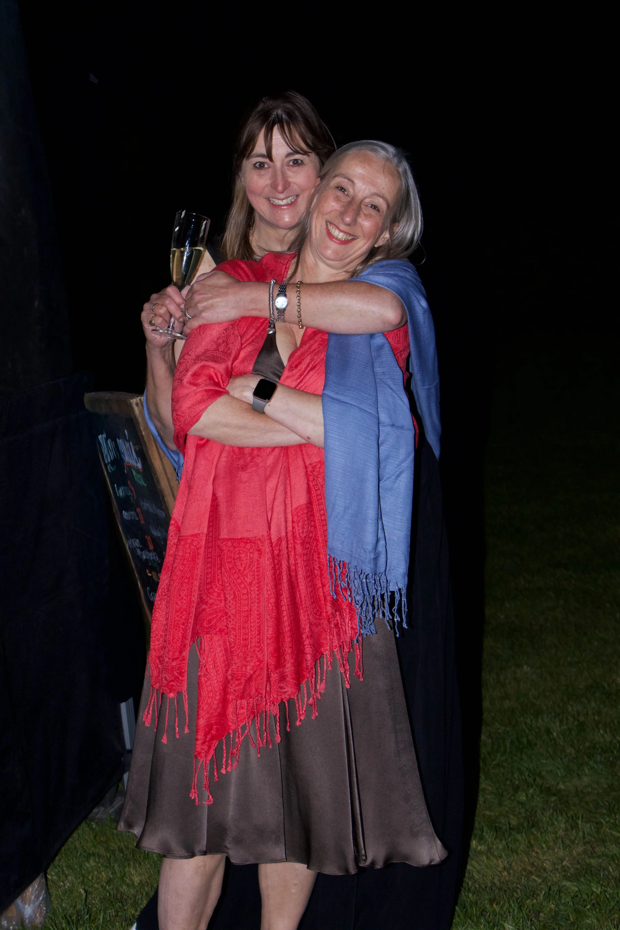 Two women smiling and hugging. The woman in the front wears a red shawl and holds a glass of champagne, the woman in the back has brown hair and wears a dark dress, an American Girl cheerleading uniform.