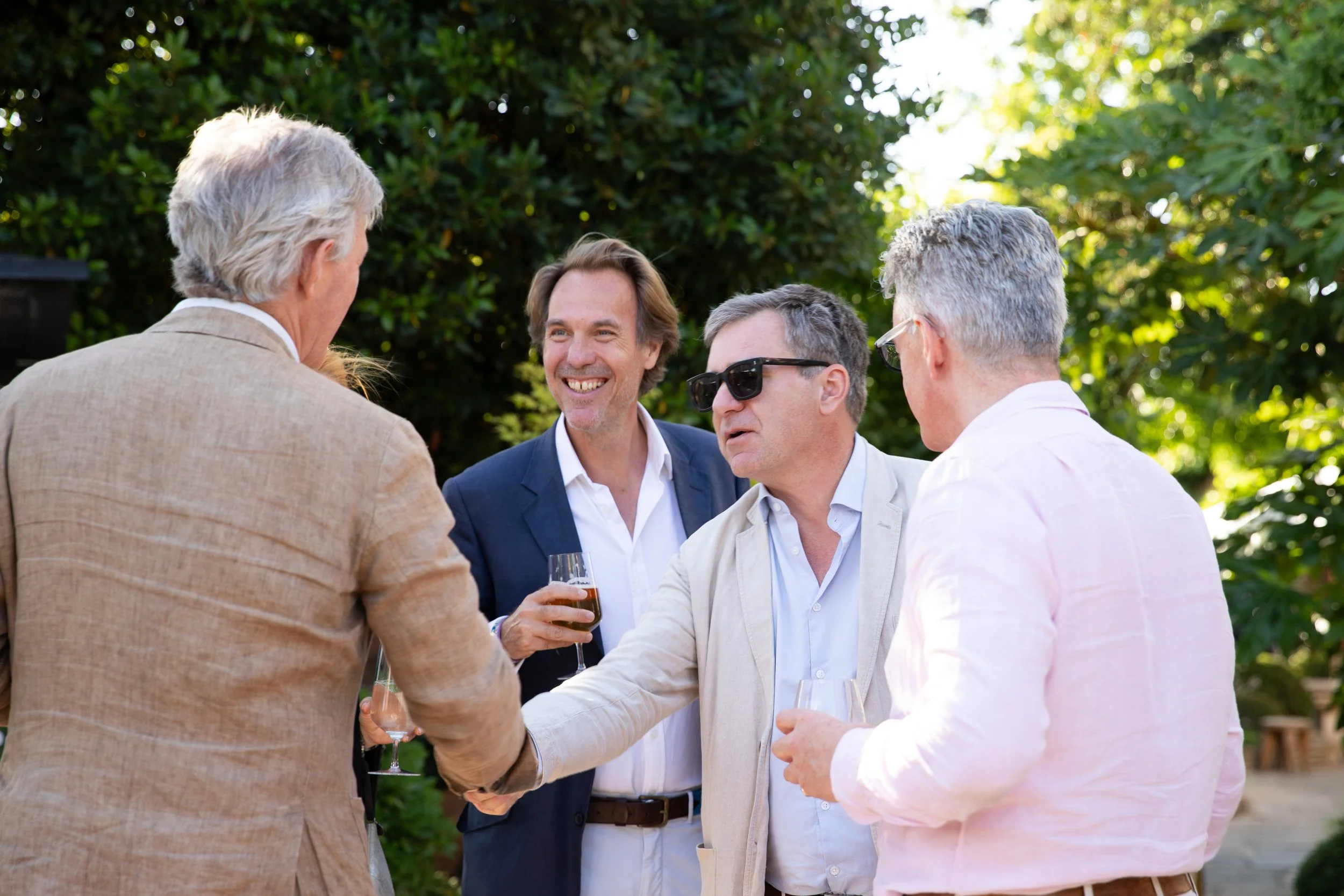 Group of five men having conversation at outdoor event, standing in a circle, holding drinks, with green foliage and sunlight in the background.