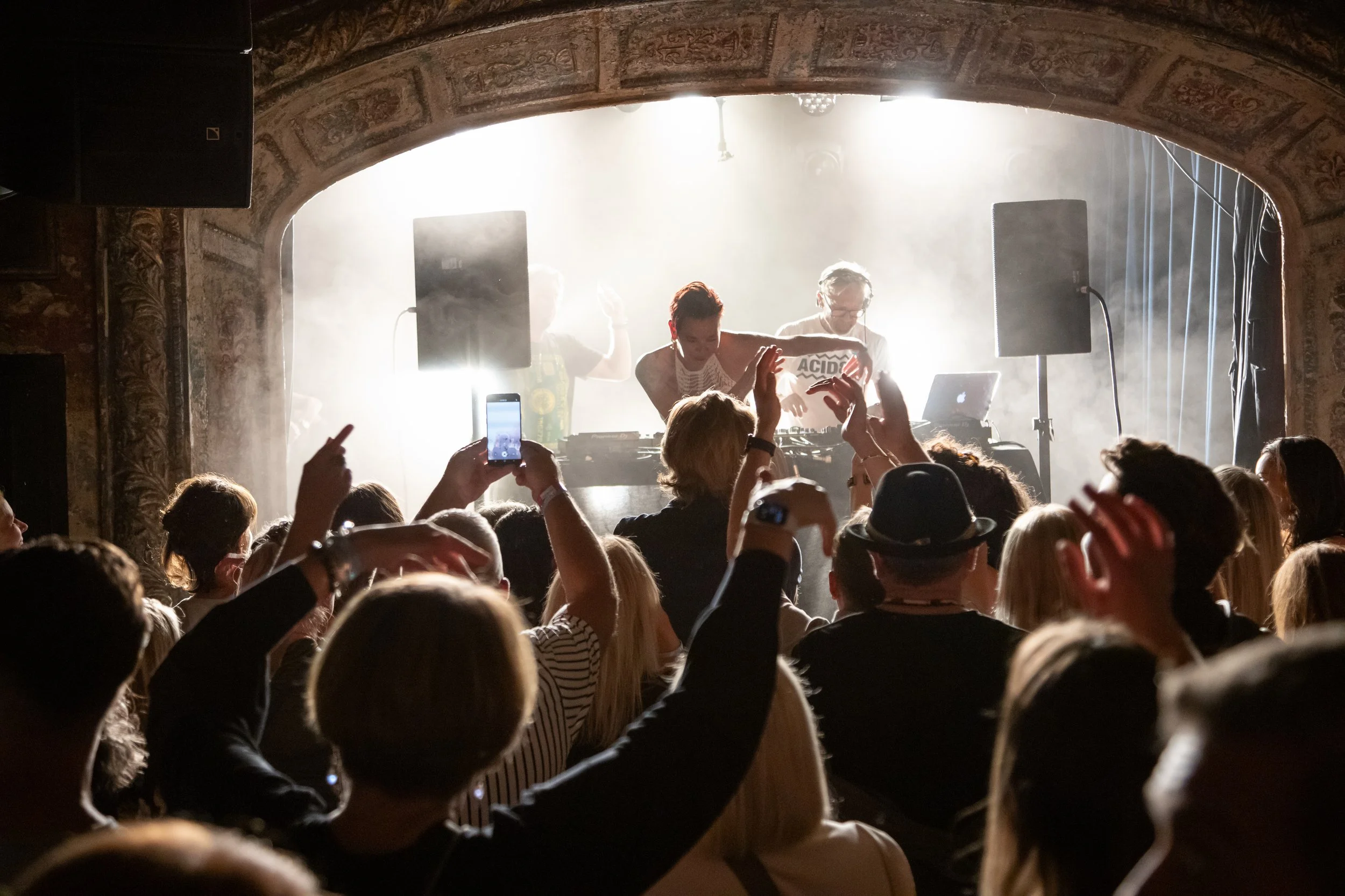 A DJ performing at a concert with a crowd enjoying the event. The stage features lighting effects and sound equipment, with some audience members capturing the moment on their phones.