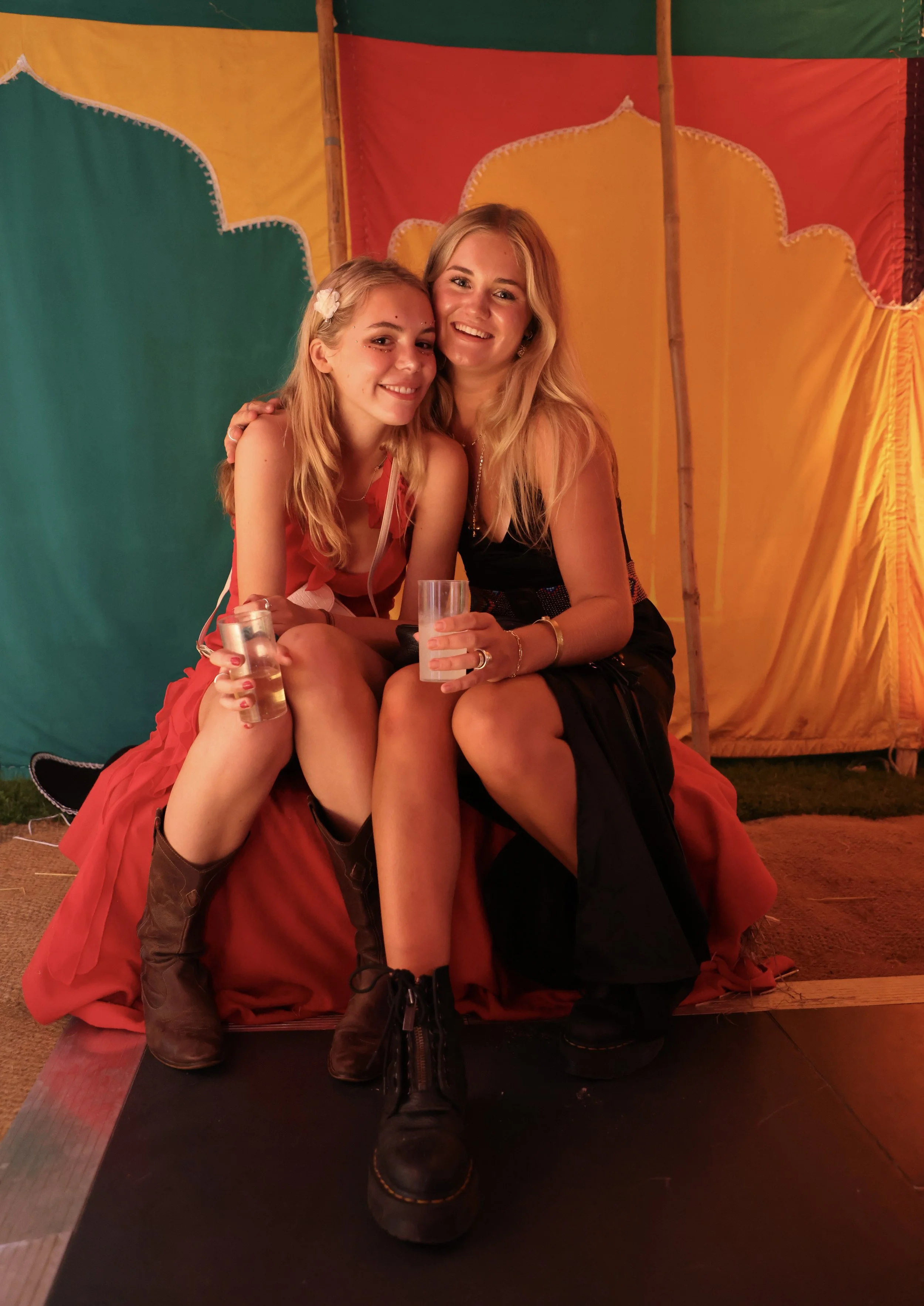 Two women sitting closely together, smiling, at a colorful carnival or circus tent with green, yellow, and red fabric backgrounds. They are holding drinks, enjoying the moment.