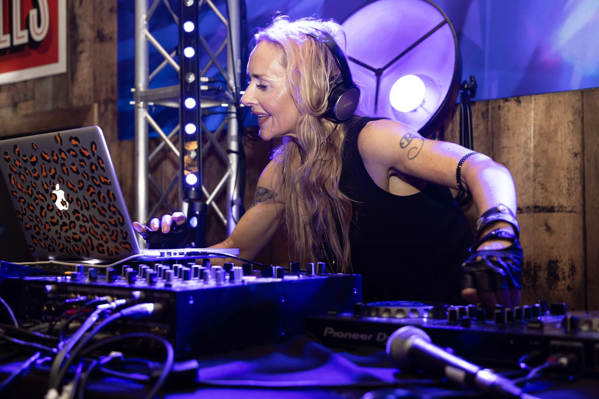 A woman with long blonde hair wearing headphones and a black tank top is DJing at a music event. She is smiling while adjusting music equipment, which includes a DJ mixer and a laptop with a leopard print cover. The background has colorful stage ligh