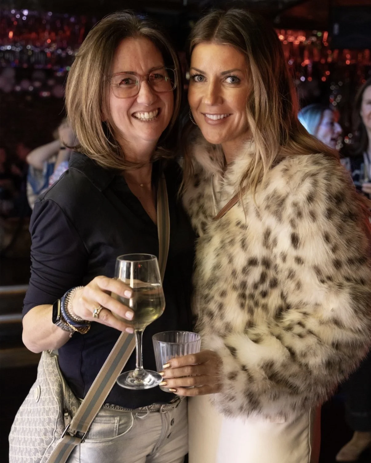 Two women posing together at an indoor party, one holding a glass of white wine, smiling at the camera.