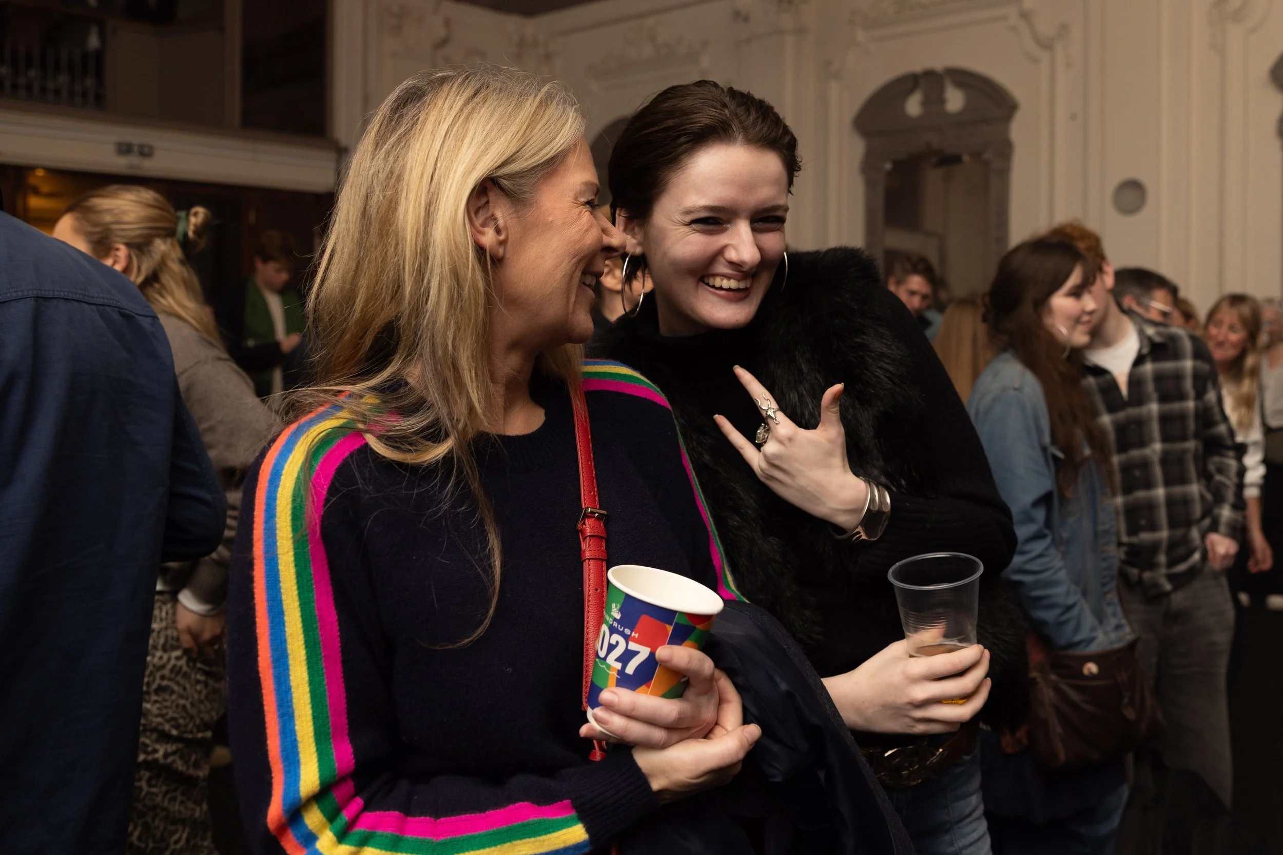 Two women smiling and chatting at an indoor social event. One woman wears a black outfit with a rainbow-striped cardigan, and the other woman wears a black outfit with hoop earrings, holding cups.