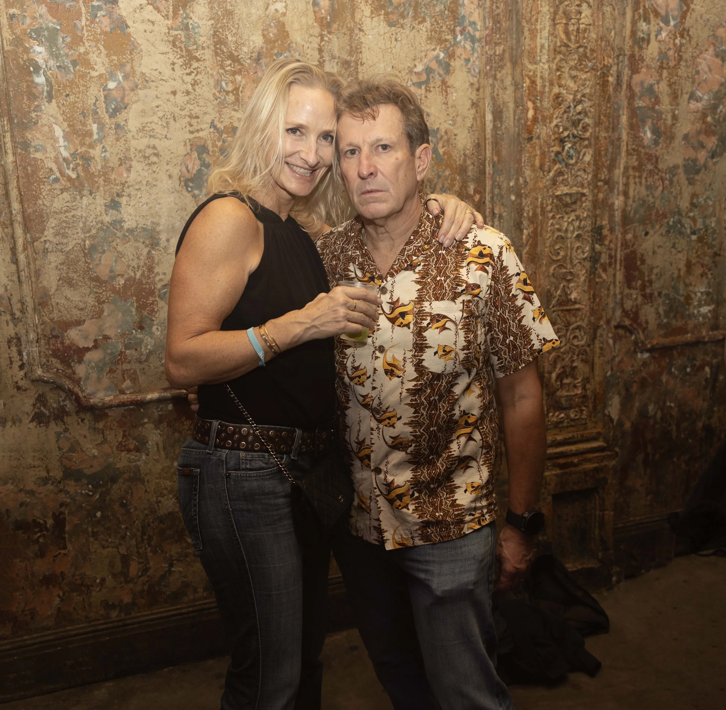 Two people, a woman with blond hair and a man with curly hair, standing close together and smiling at the camera against a textured, weathered wall.