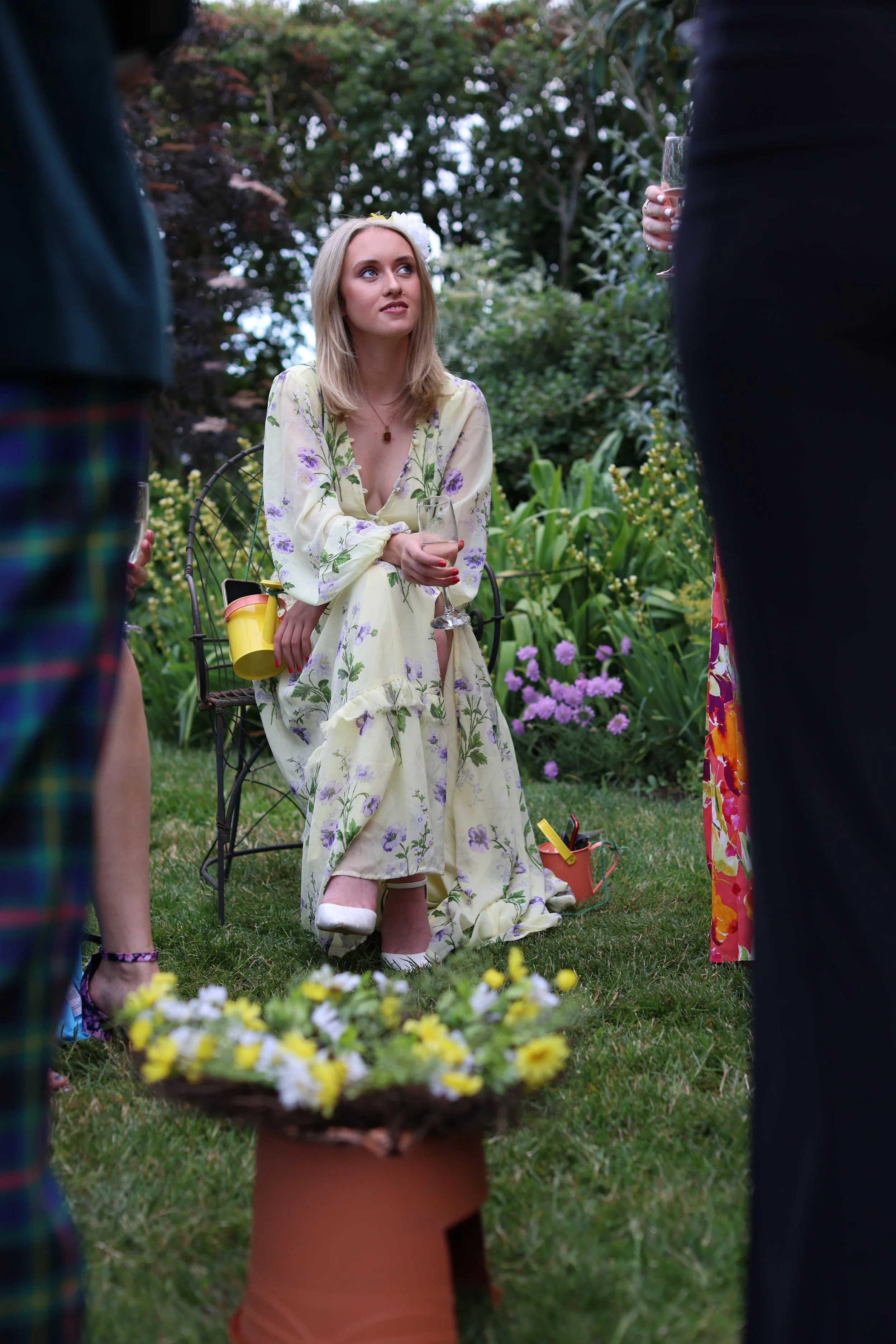 A woman in a floral yellow dress sitting on a garden chair, holding a glass of wine, attending an outdoor gathering. Several people are around her, some holding drinks, in a lush garden setting with flowers and greenery.