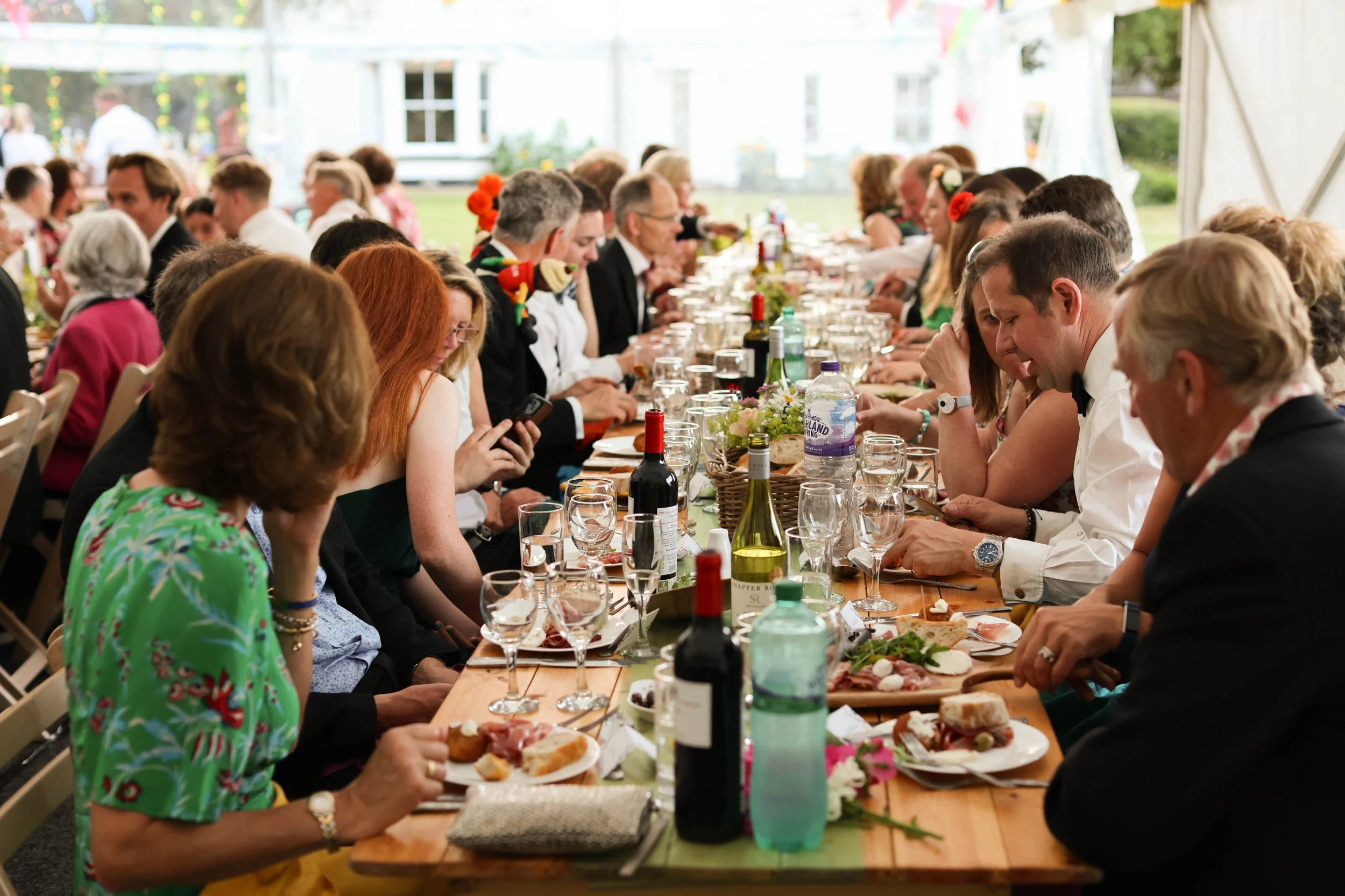 People seated at a long table outdoors, enjoying a meal and drinks, at a festive gathering or celebration.