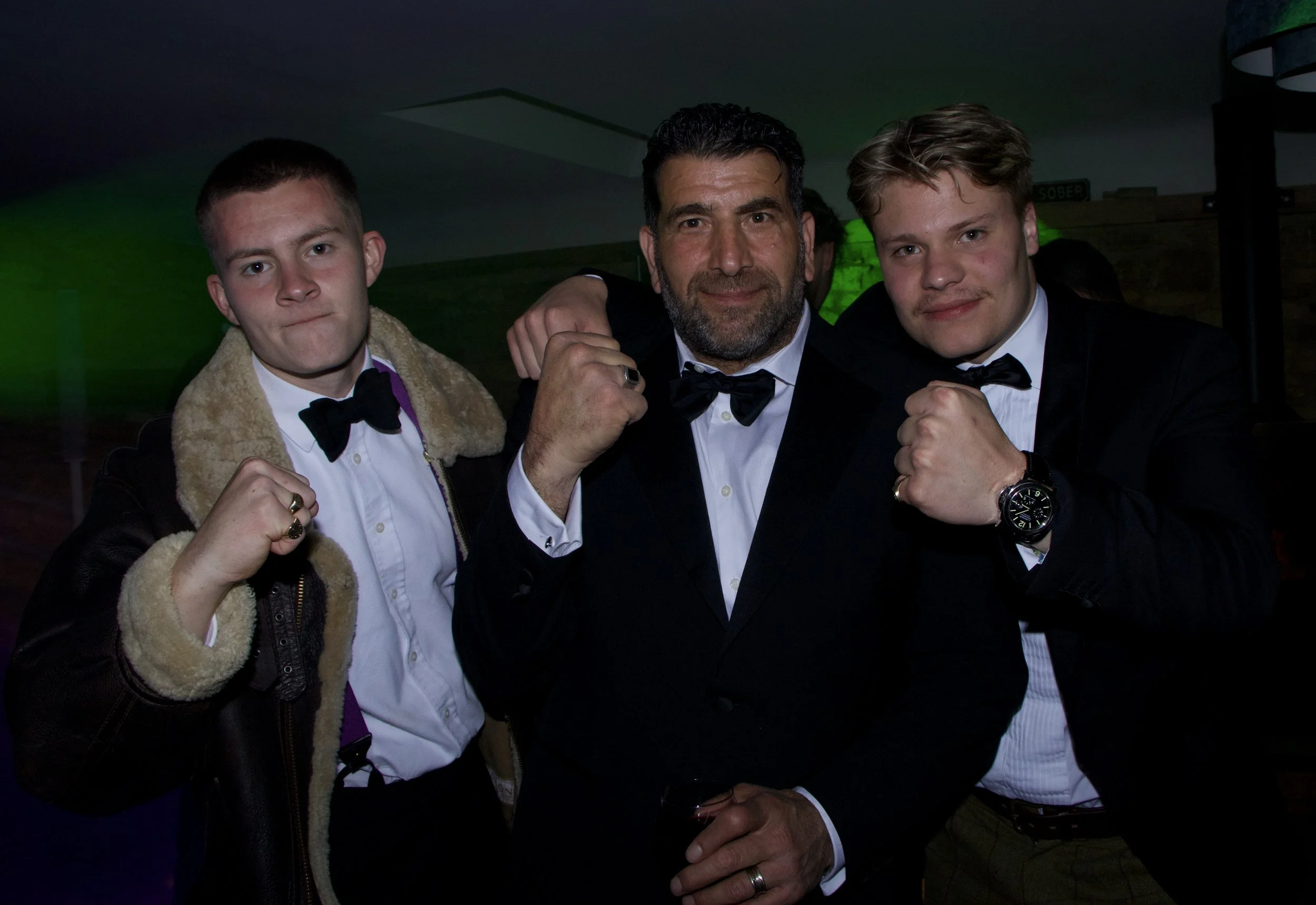 Three men dressed in tuxedos with bow ties posing together in a dark indoor setting, making fists and smiling.