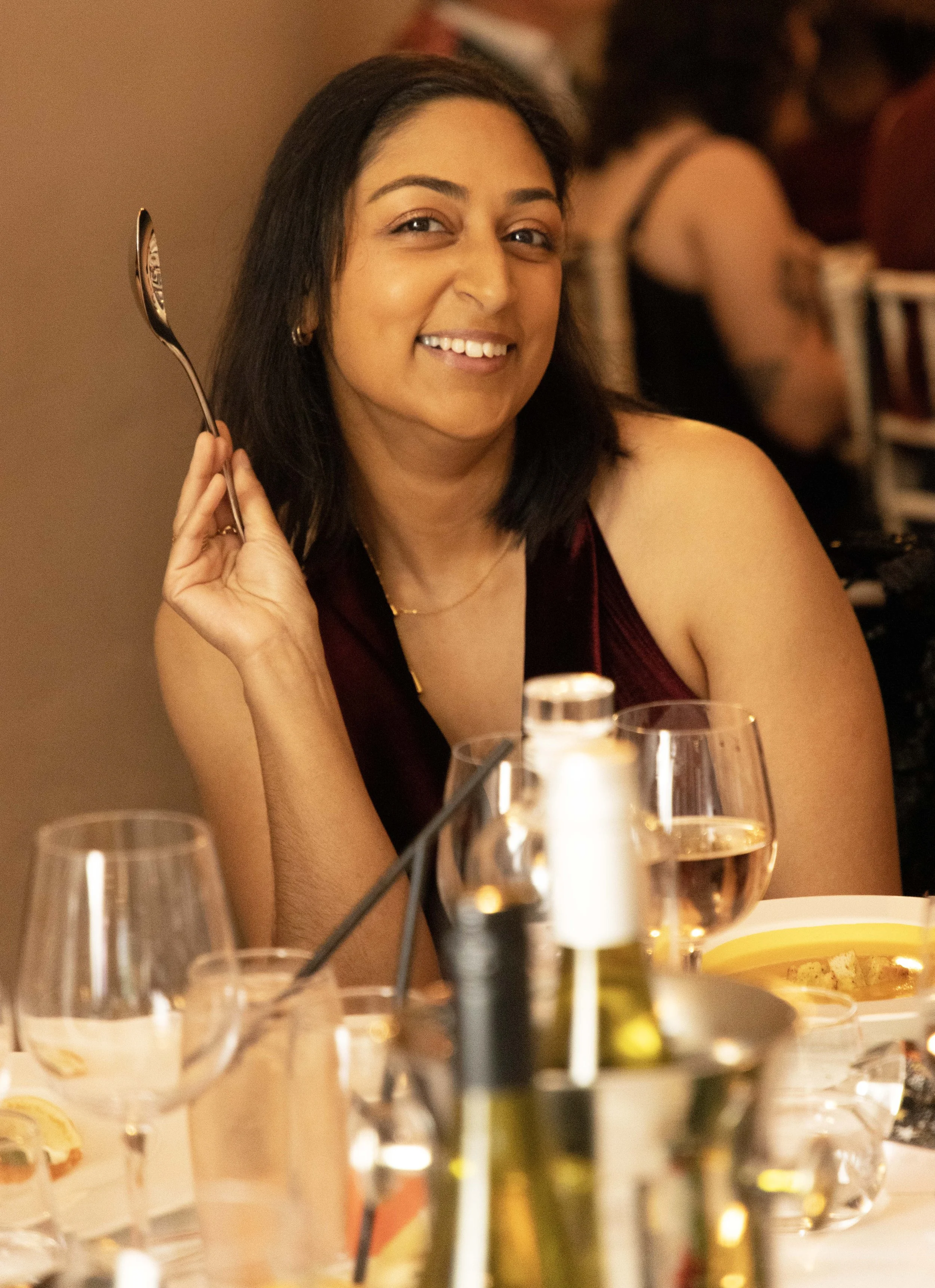 A woman with shoulder-length dark hair, smiling and holding a spoon, sitting at a table set with wine glasses, plates, and bottles of oil or vinegar, during a dinner gathering in a warmly lit setting.