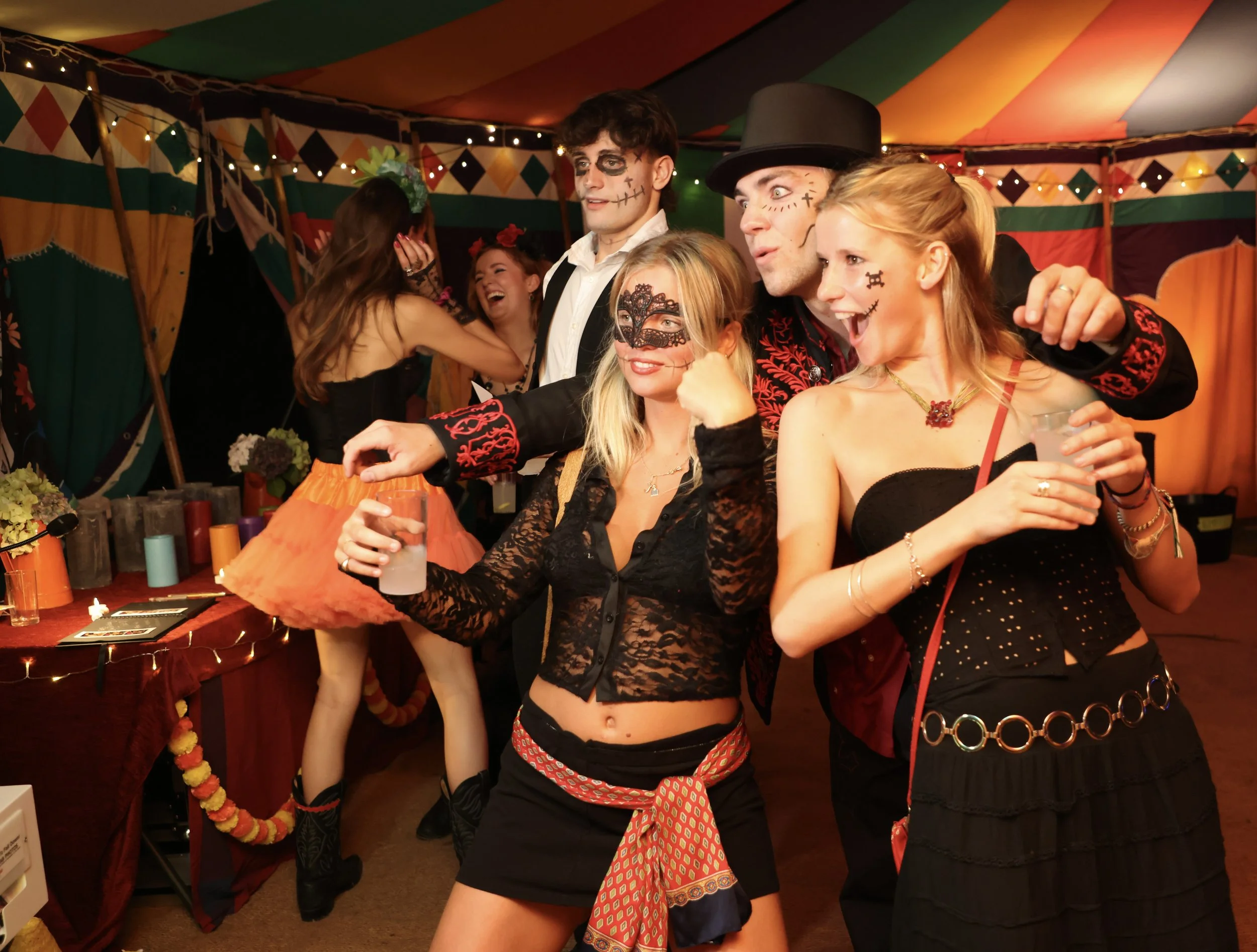 Group of people at a costume party, wearing Halloween-themed costumes with face paint, inside a decorated tent with colorful drapes and string lights.