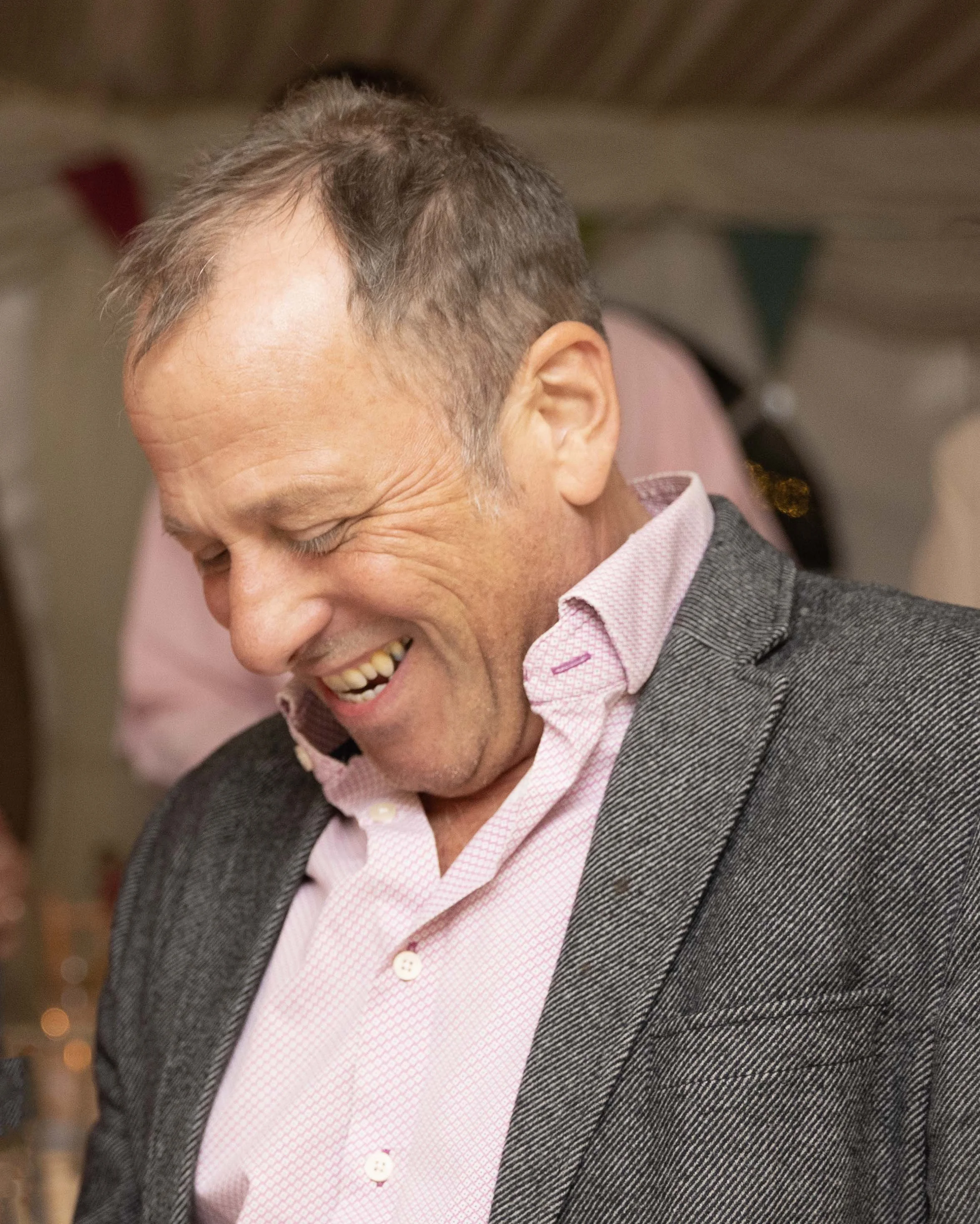 A man with short brown hair, smiling and looking downward, wearing a pink checkered shirt and a gray suit jacket, indoors with blurred decorations in the background.