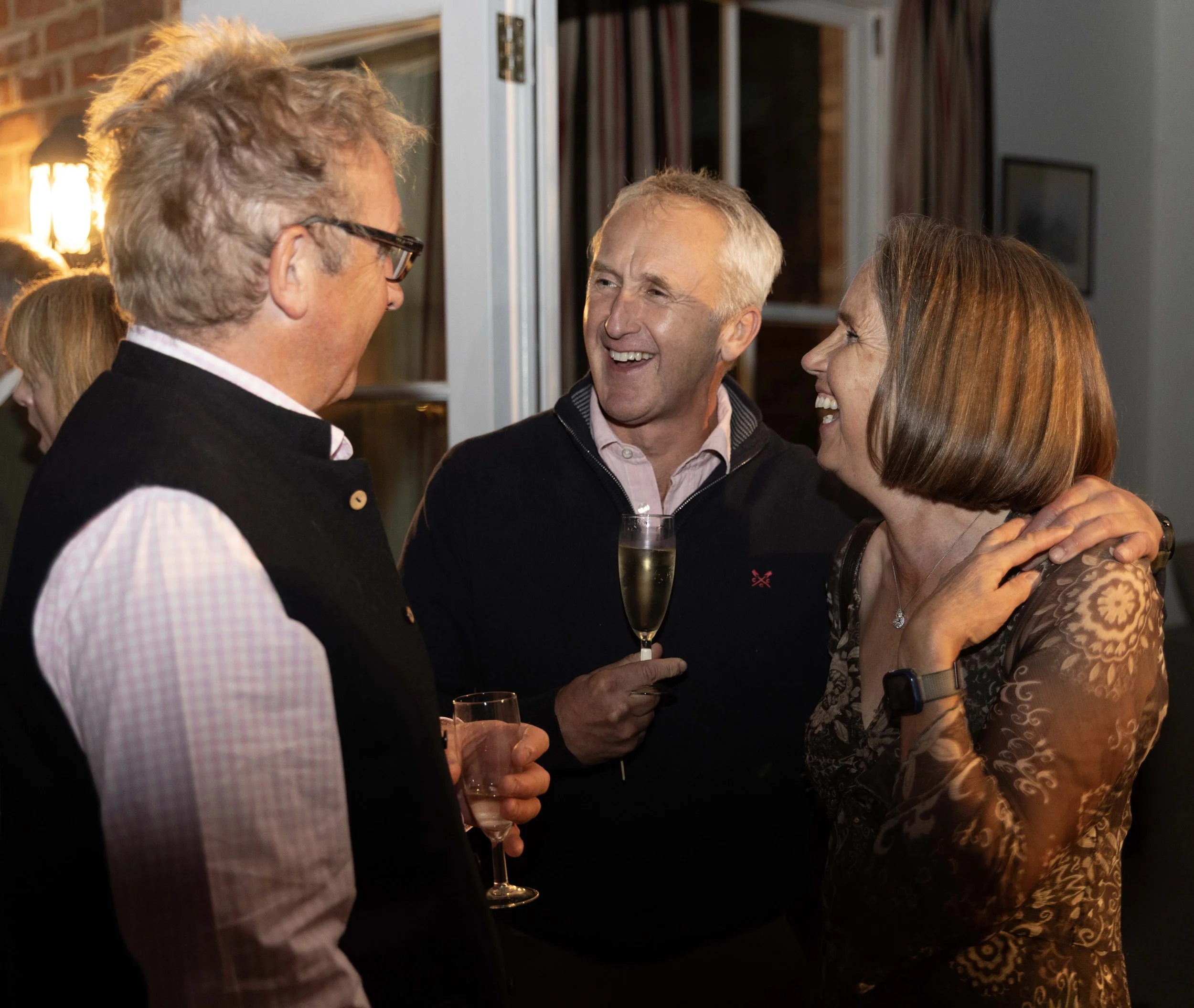 Three people smiling and chatting at a social gathering, holding drinks, in a warmly lit indoor setting.