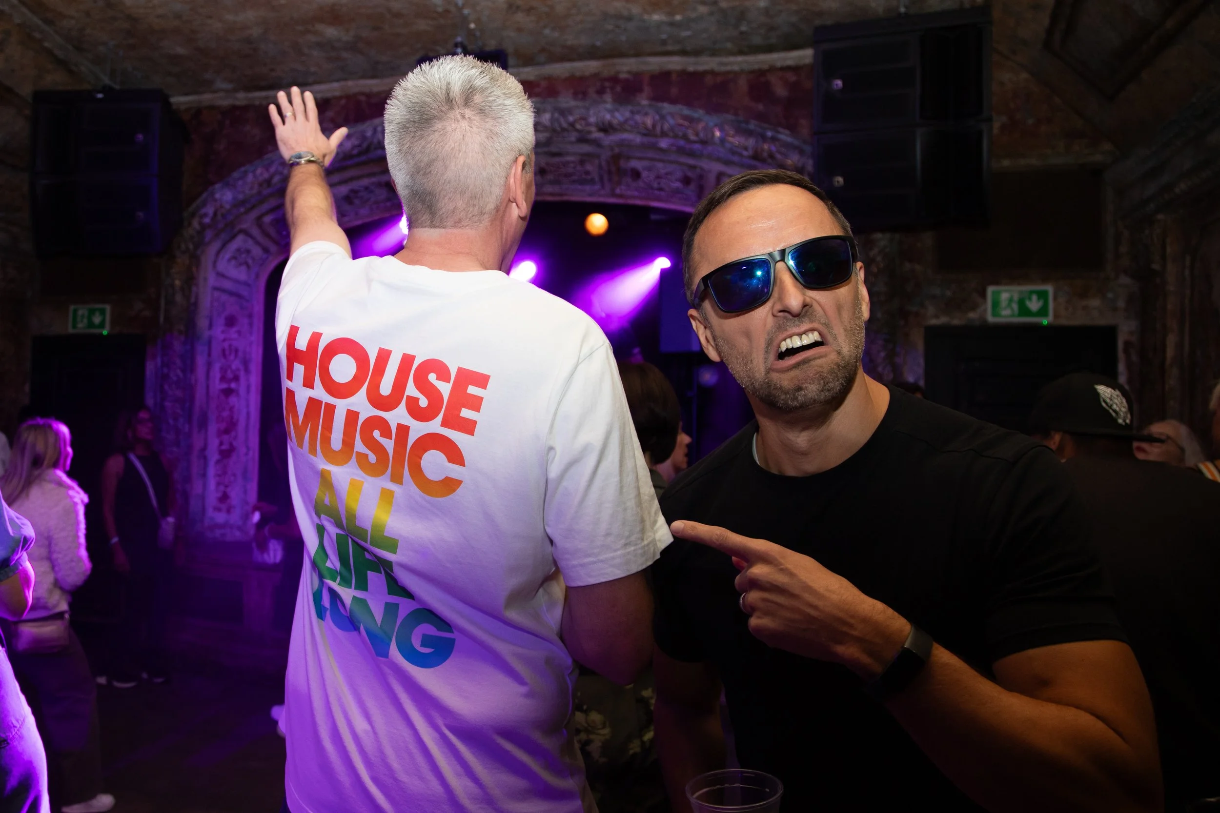 Two men at a lively indoor concert or event. The man on the left has short, light-colored hair and is wearing a white T-shirt with colorful text reading 'HOUSE MUSIC ALL NIGHT.' The man on the right has short dark hair, sunglasses, and is making a fu