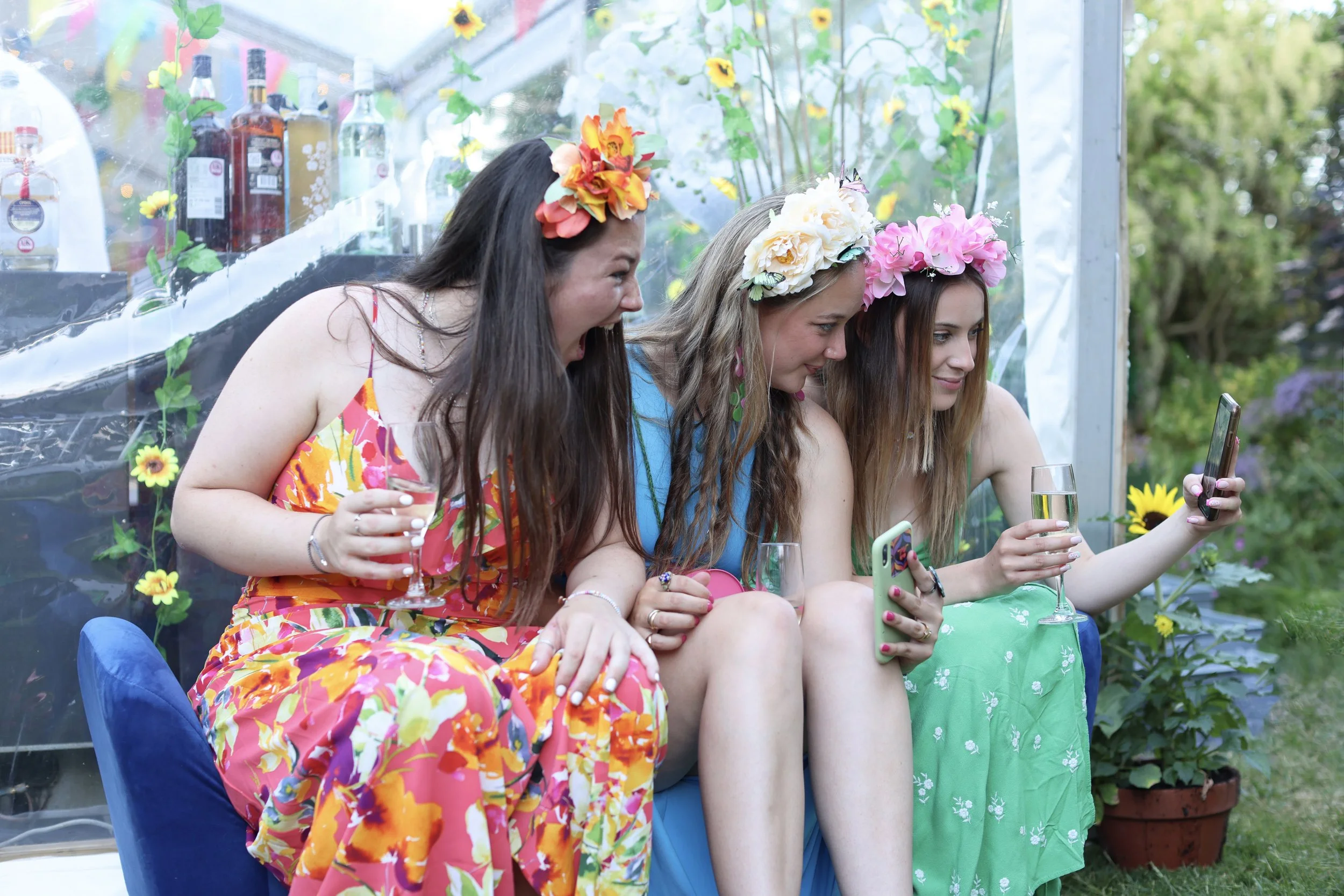 A group of three women sitting together, wearing colorful dresses and flower crowns, outdoors with drinks in hand, taking selfies or photos with their phones.