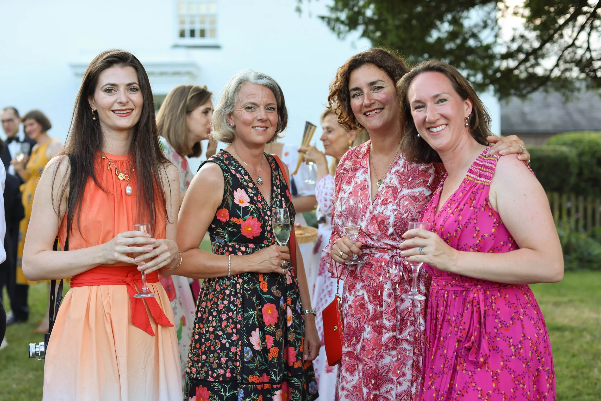 Four women in colorful dresses smiling and holding glasses of wine at an outdoor gathering, with some guests and trees in the background.