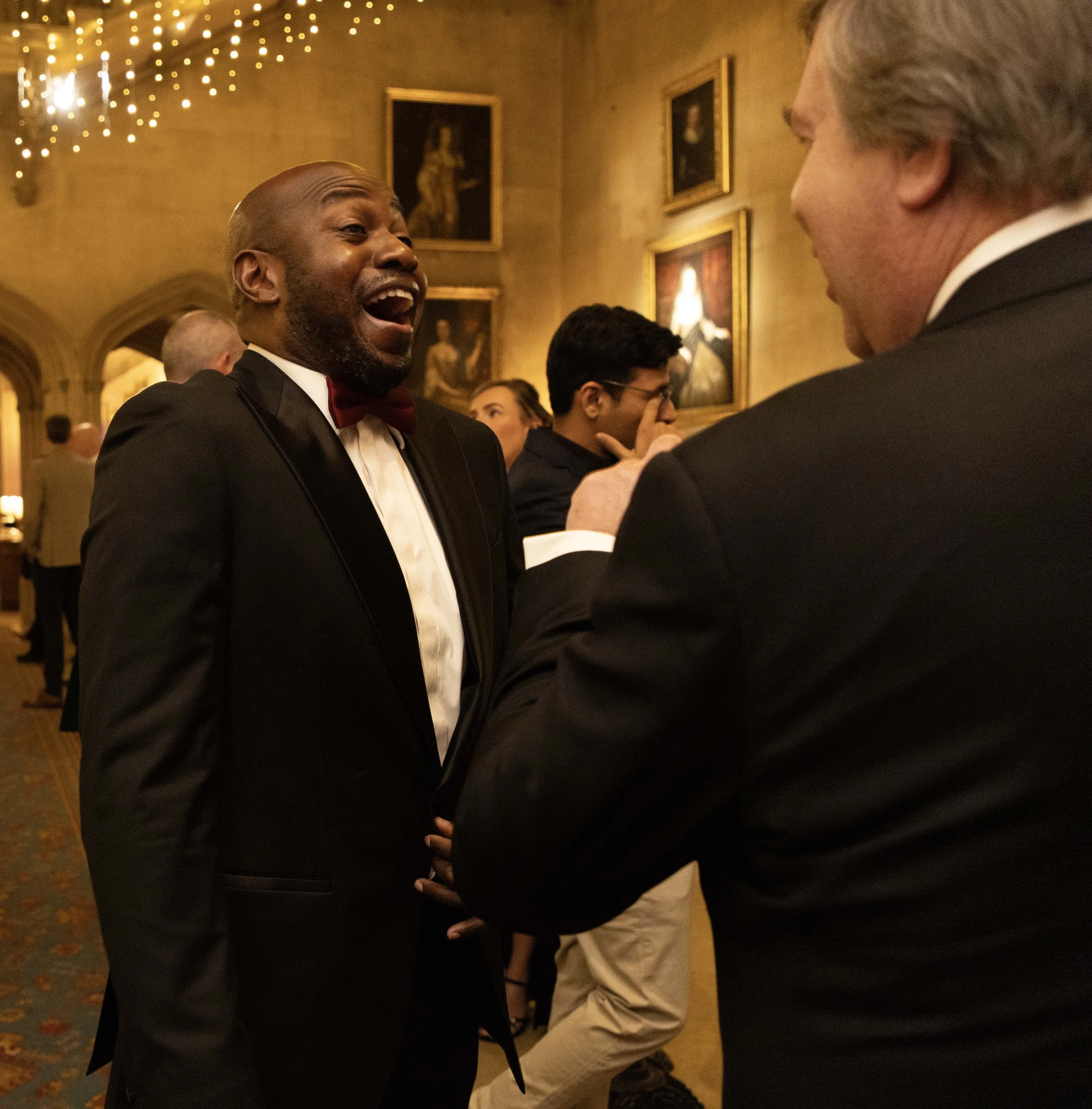Two men dressed in formal tuxedos engaging in conversation at an event in a warmly lit room with framed paintings on the wall.