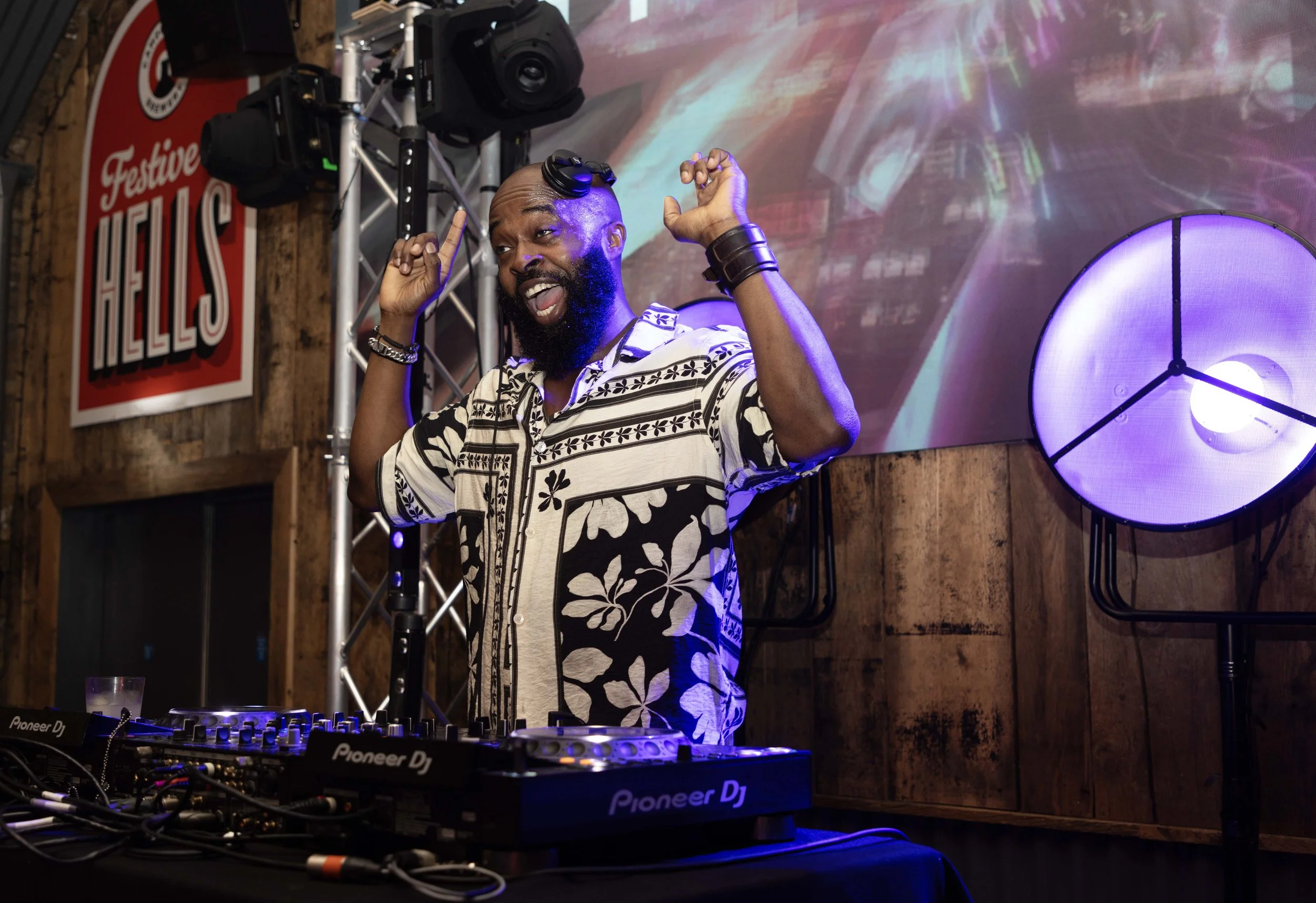 A DJ with a beard and a patterned shirt is performing at a music event. He is using turntables and a mixer, with his hands raised and expressive facial expression. The background features a large digital screen and a purple-lit wall decoration. The s