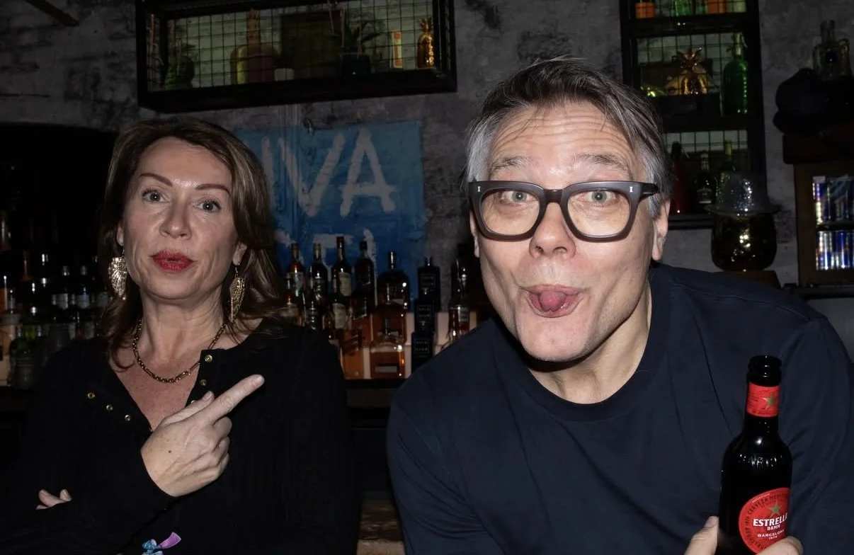 A man and woman posing for a photo in a bar. The woman has shoulder-length brown hair, is wearing gold earrings, and is pointing at the man. The man has gray hair, glasses, and is holding a beer bottle with a red label. The background shows shelves w