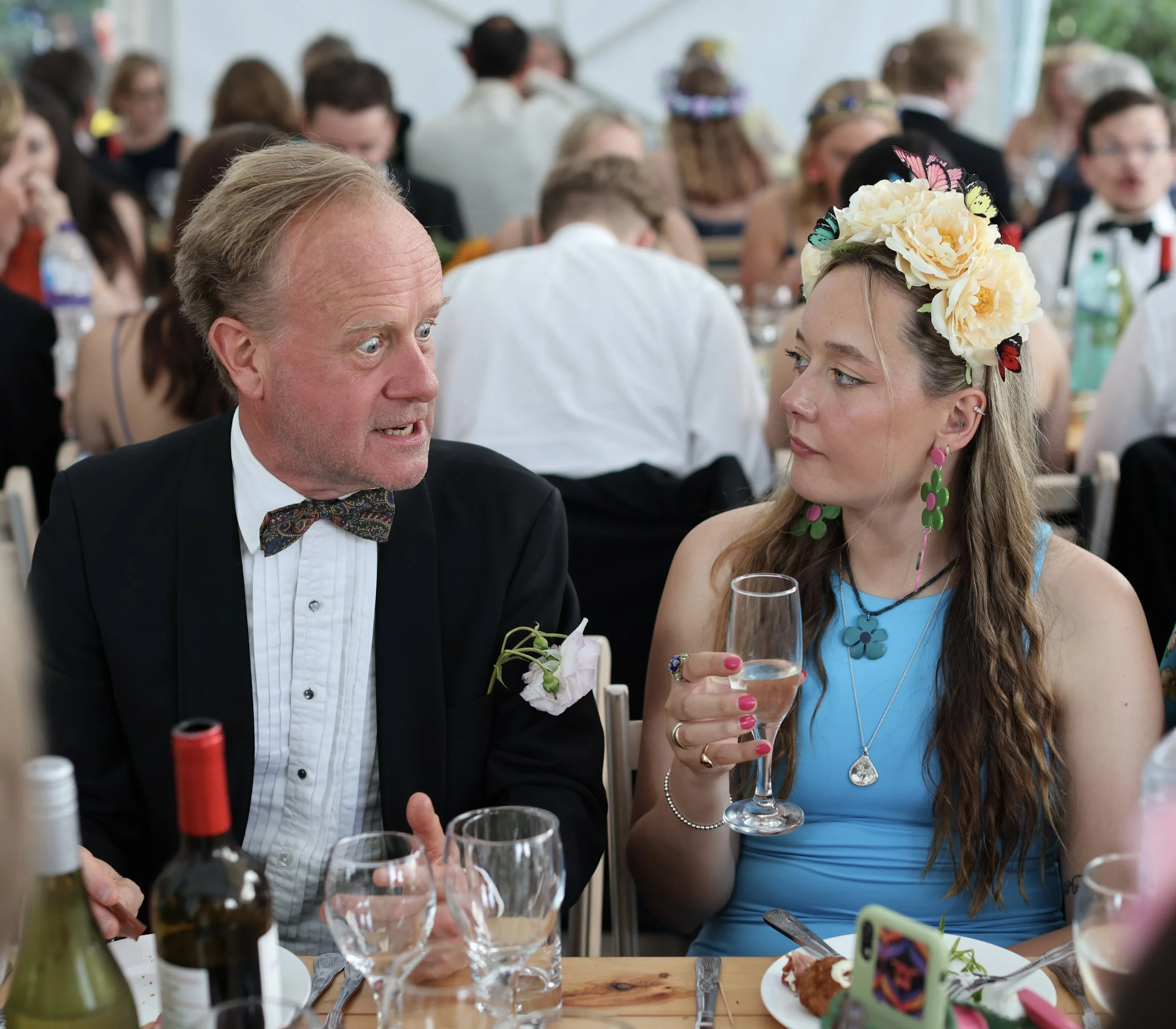 A man and a woman dressed for a formal event, possibly a wedding, are sitting at a table engaged in conversation. The woman is wearing a blue dress and floral earrings, with a floral headpiece decorated with butterflies. The man is wearing a tuxedo w