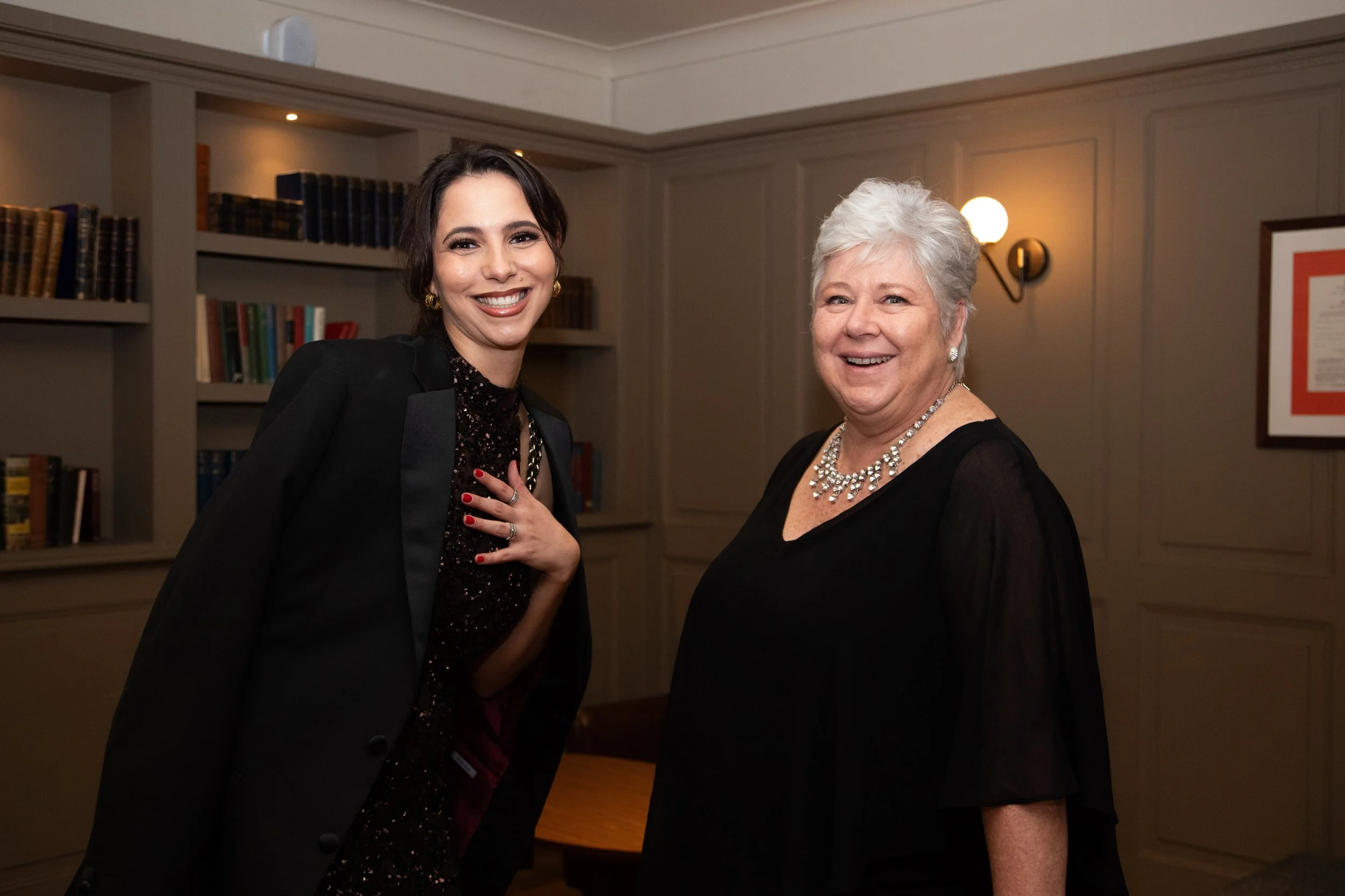 Two women smiling indoors, one with dark hair and dressed in black, the other with gray hair and wearing a black dress with pearl jewelry, standing in front of bookshelves and wall decor.