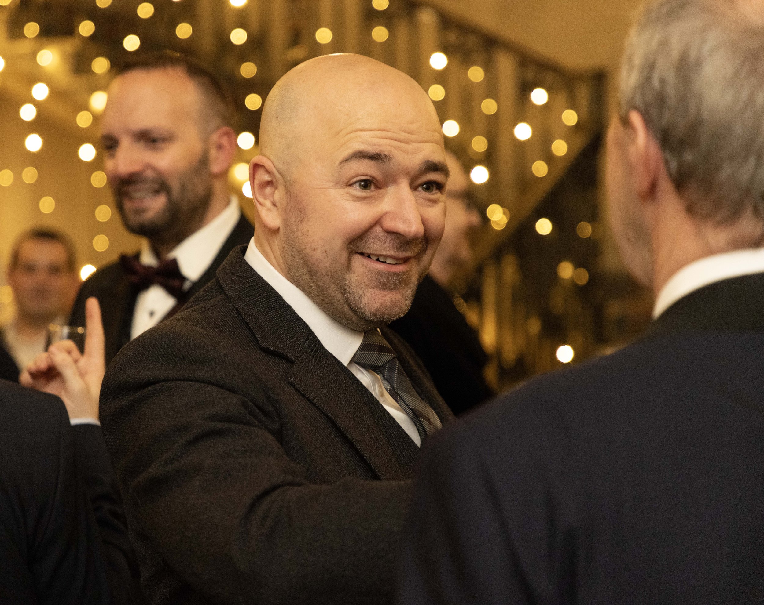 A group of men dressed in tuxedos at a formal event, engaging in conversation, with blurred warm lighting and hanging lights in the background.