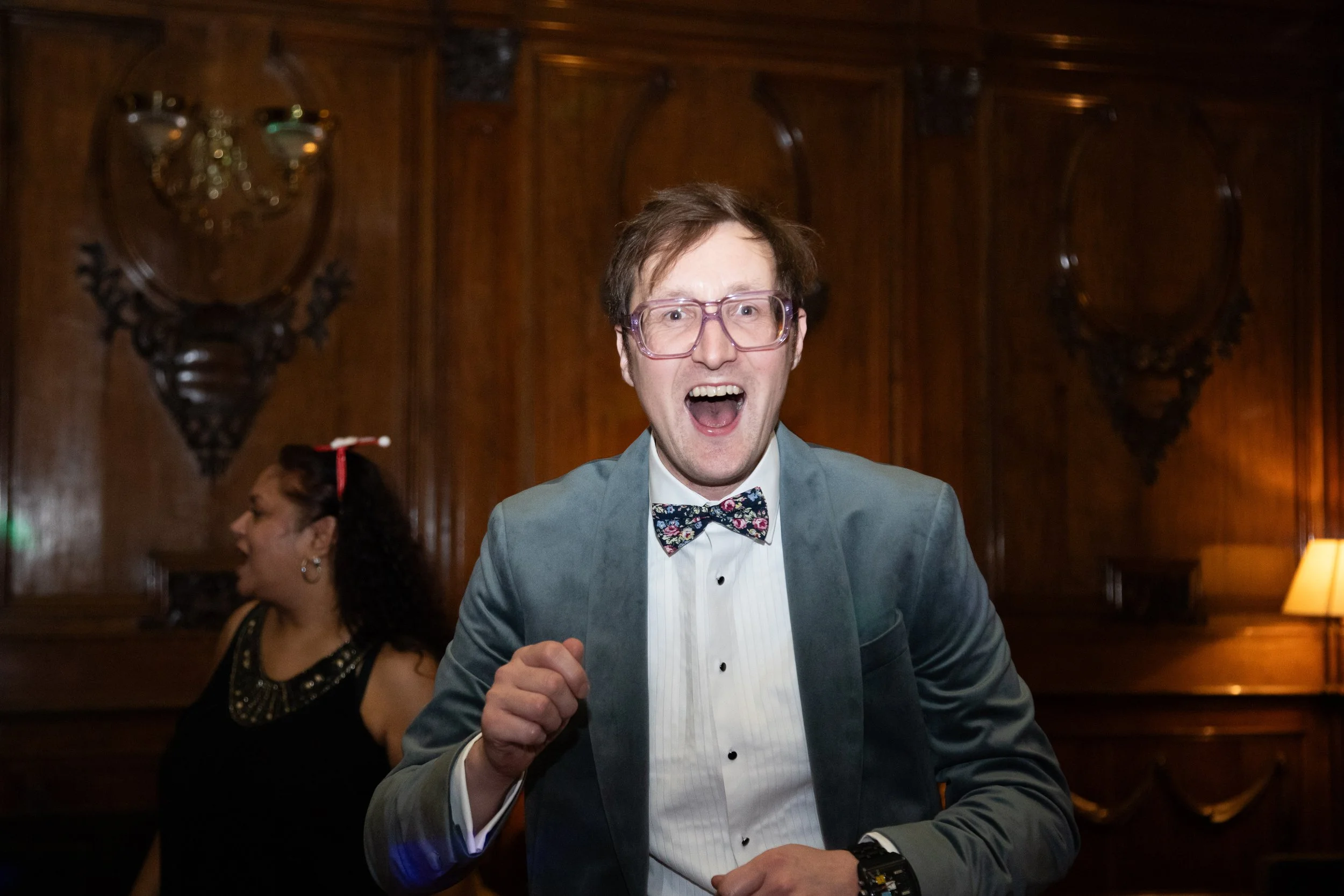 A man with glasses and a floral bow tie, dressed in a suit, is smiling and raising his fist, in a dimly lit room with wooden walls. A woman with earrings and a black dress is partially visible on the left.