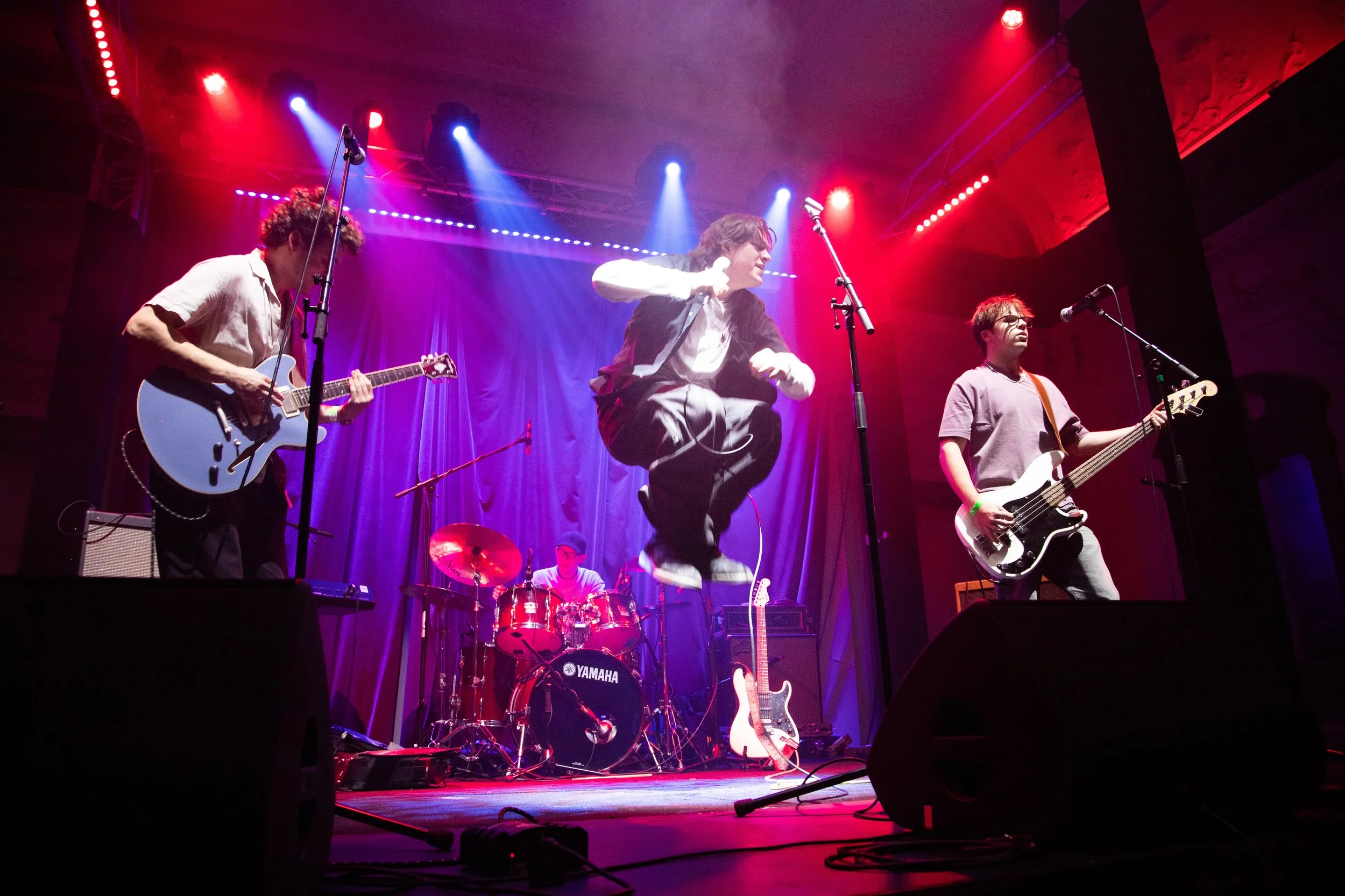 A band performing live on stage with colorful lighting, including guitarists, a vocalist, and a drummer.