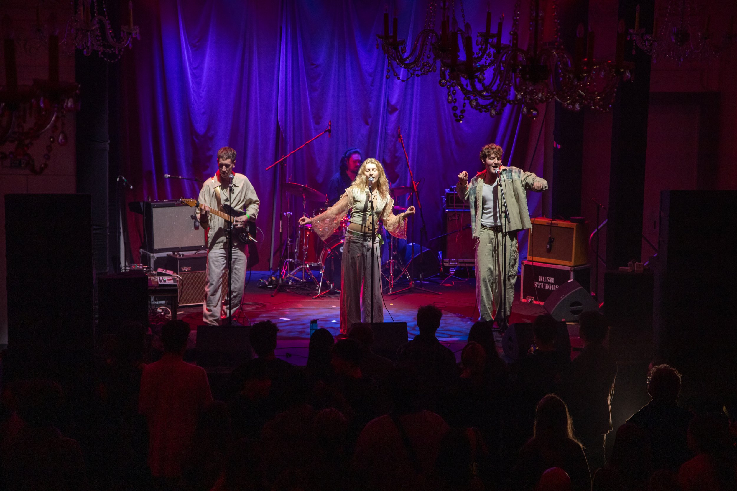 A band performs on stage with a singer and guitarist in front of a draped purple curtain, illuminated by stage lighting and chandeliers.