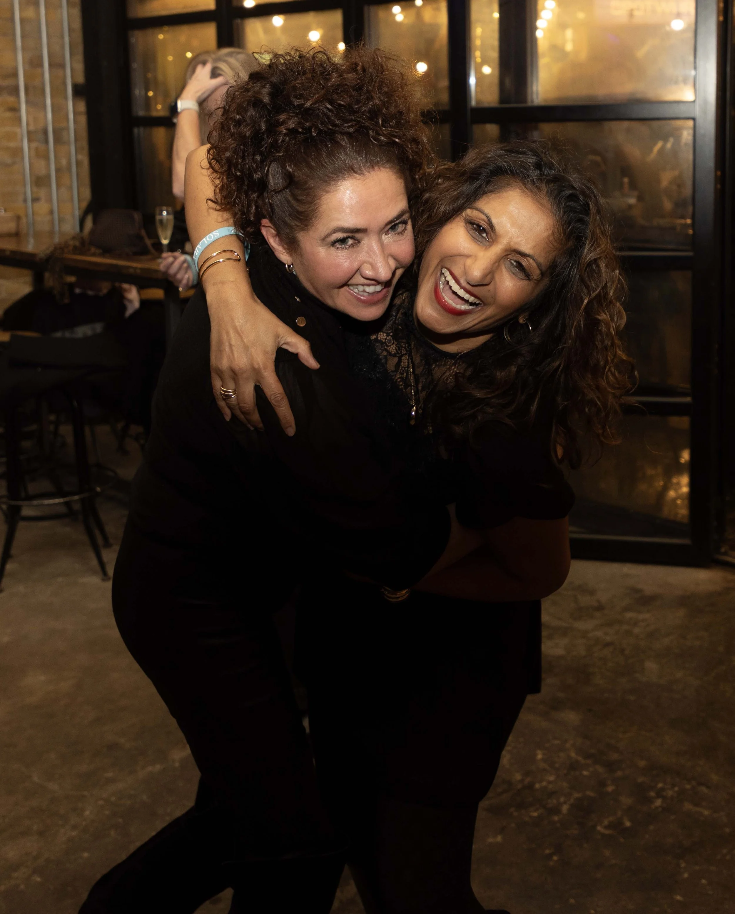 Two women hugging at an indoor gathering, smiling and showing joy, with warm lighting and glass windows in the background.