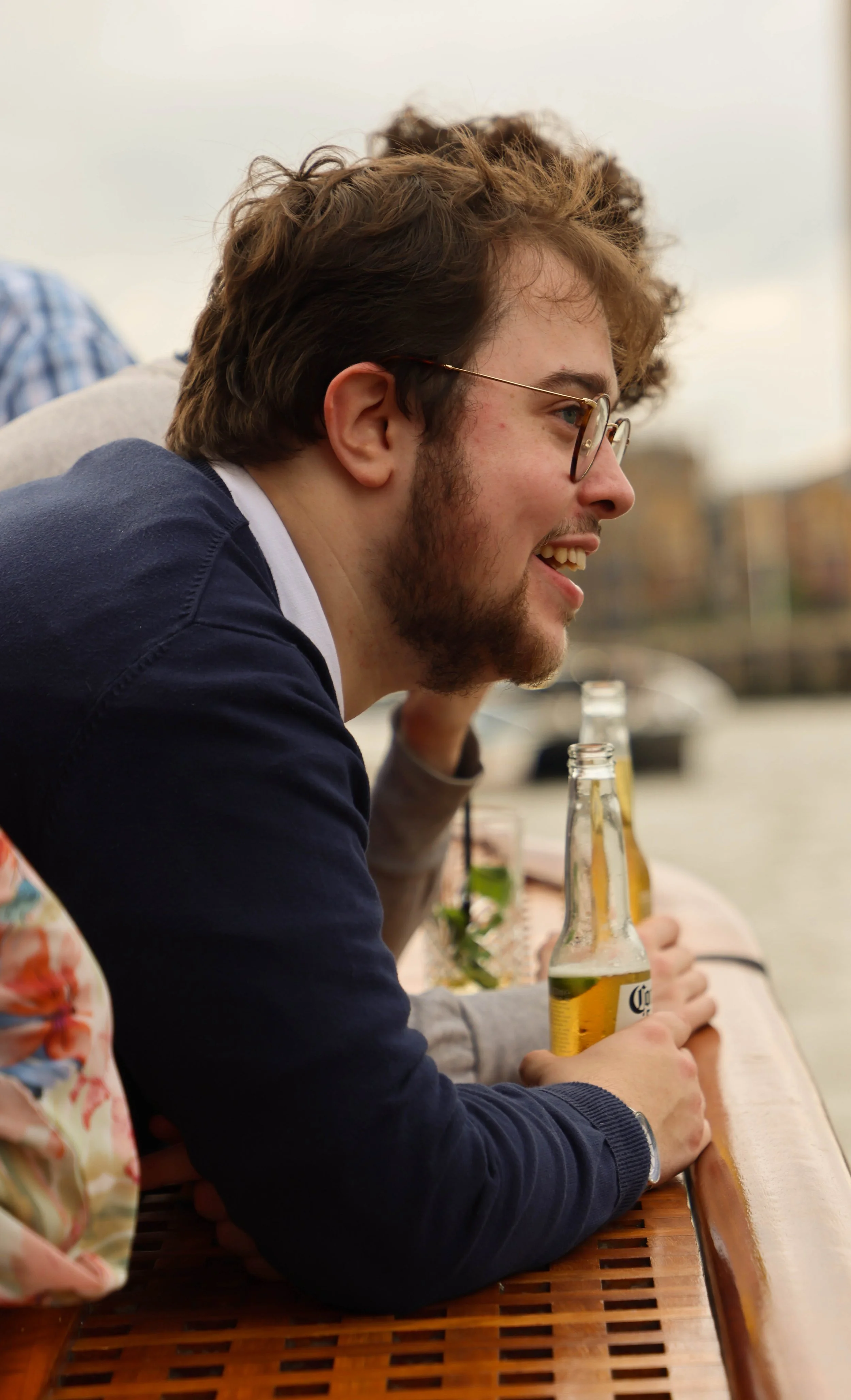 Young man with glasses and brown hair smiling while sitting at an outdoor table with drinks, during daytime.