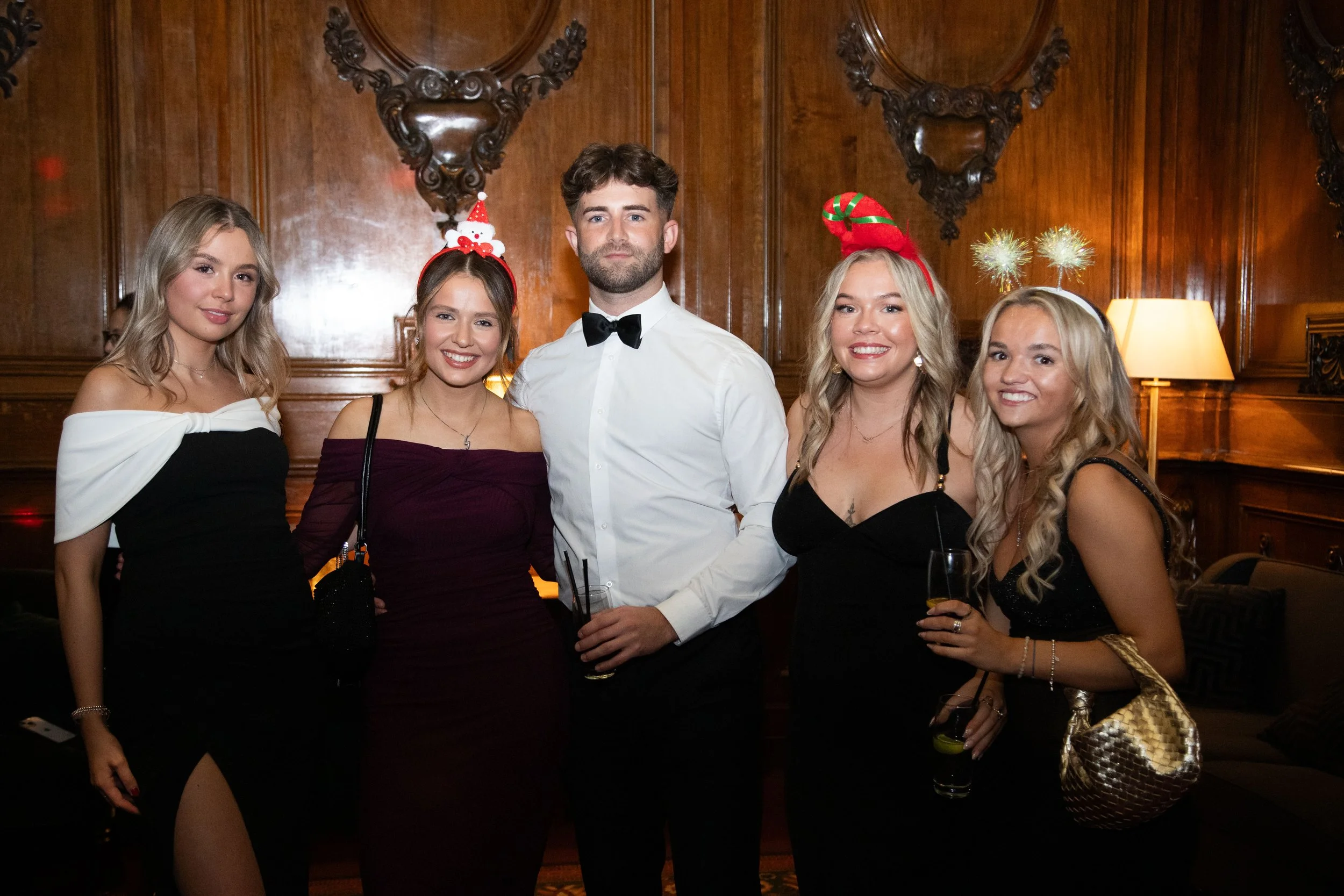 A group of five young adults dressed in formal attire at a party, with three women wearing holiday-themed headbands and a man in a white shirt with a bow tie, standing in a wood-paneled room with ornate wall decorations and lamps.