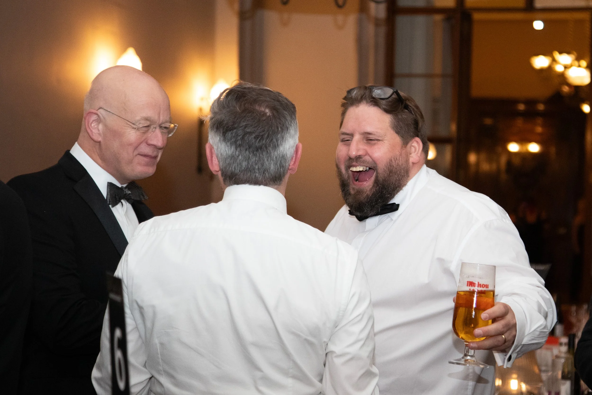Group of four men engaging in conversation at a social gathering, one holding a glass of beer. They are dressed in formal attire, including tuxedos and white shirts, in an indoor setting with warm lighting.