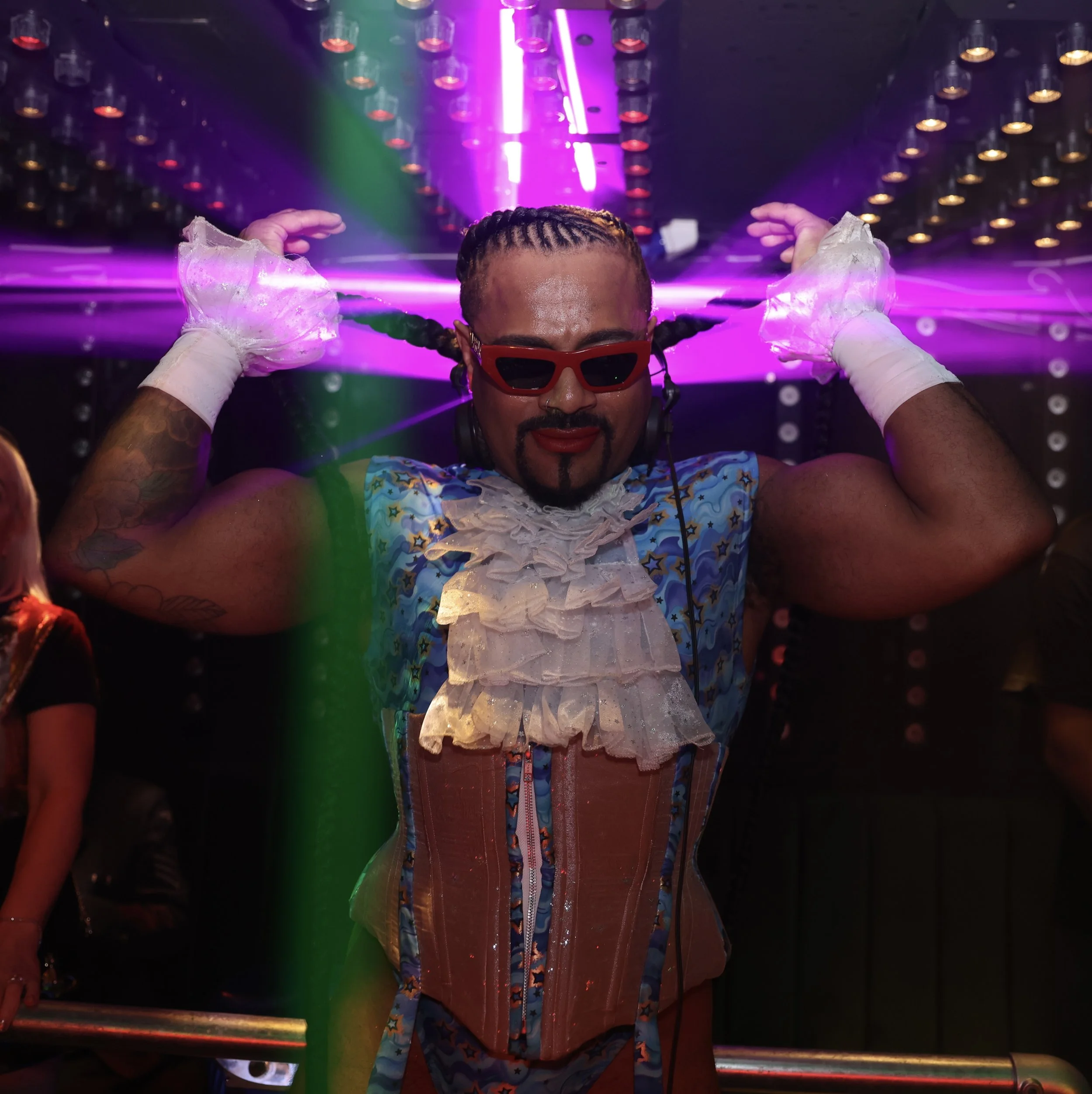 A person with braided hair wearing red sunglasses and a colorful shirt in a space with vibrant purple, green, and pink neon lights, surrounded by numerous small lights on the ceiling.