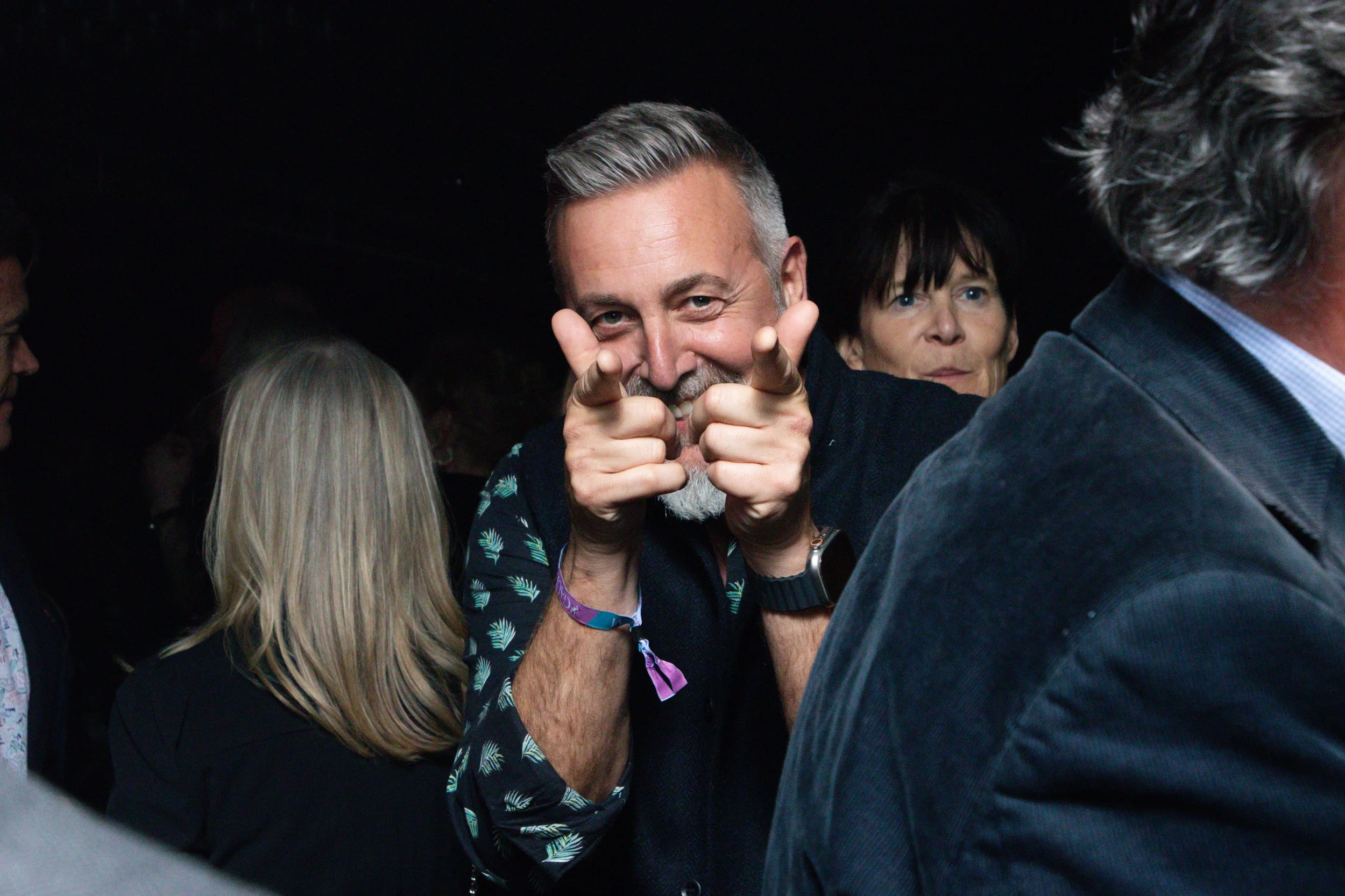A man with gray hair and a beard making a playful pointing gesture at the camera, surrounded by other people.