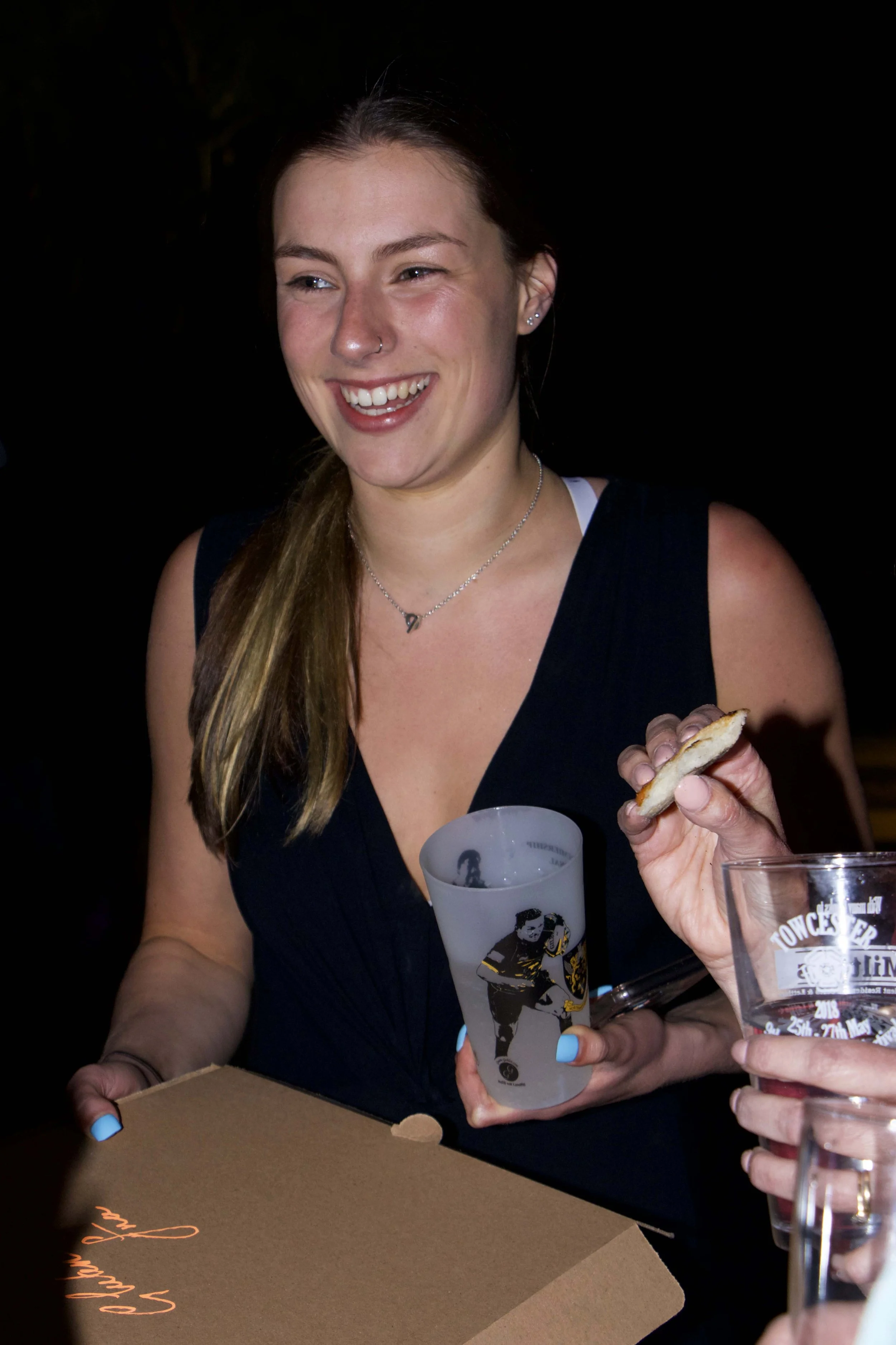 A young woman with long brown hair and a nose ring smiling while holding a tall, frosted glass with a racing car design on it. She is wearing a navy blue sleeveless top and a star-shaped necklace. People are holding drinks in clear glasses around her