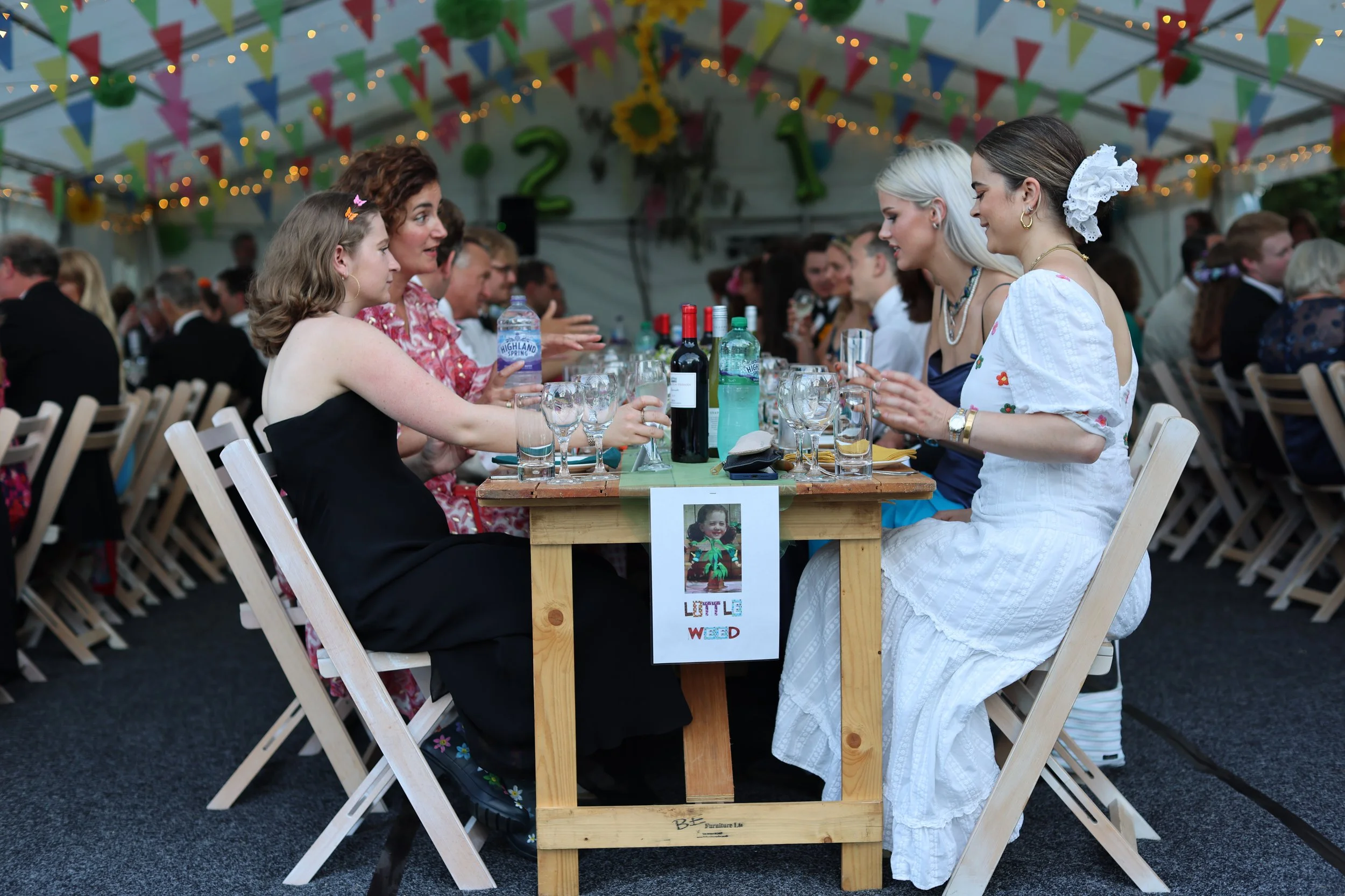 A group of people sitting around a dining table under a decorated tent celebrating a gathering or party. The table has glasses, wine bottles, and water bottles, with colorful banners and lights hanging above. There is a photo of a child on the front 
