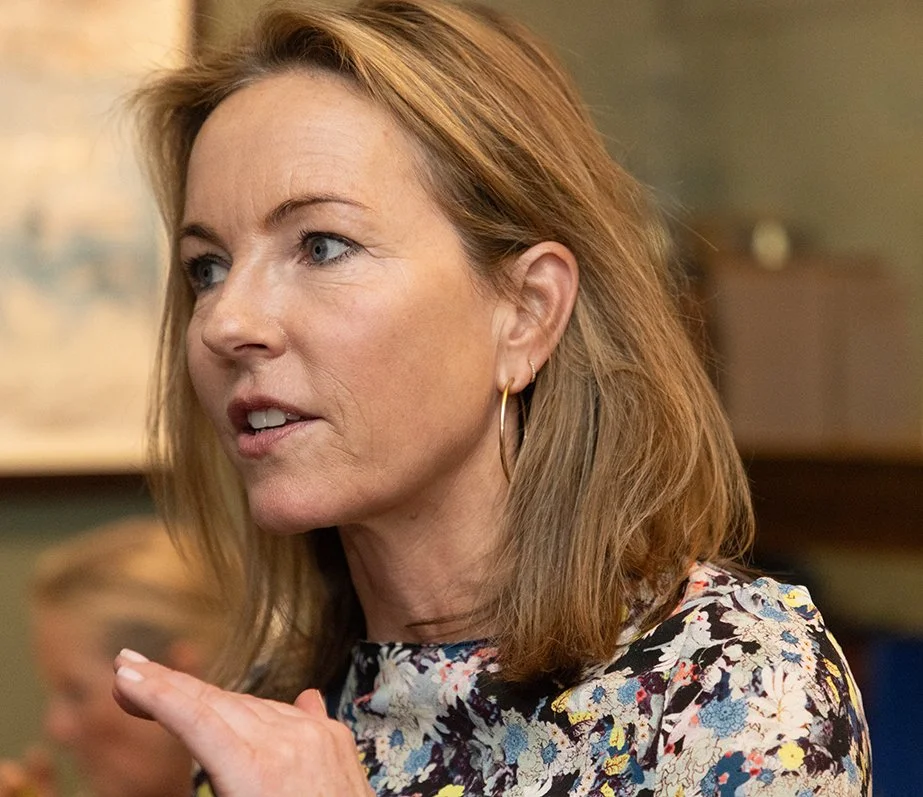 A woman with light brown hair and blue eyes, wearing earrings and a floral top, speaking or explaining something indoors.