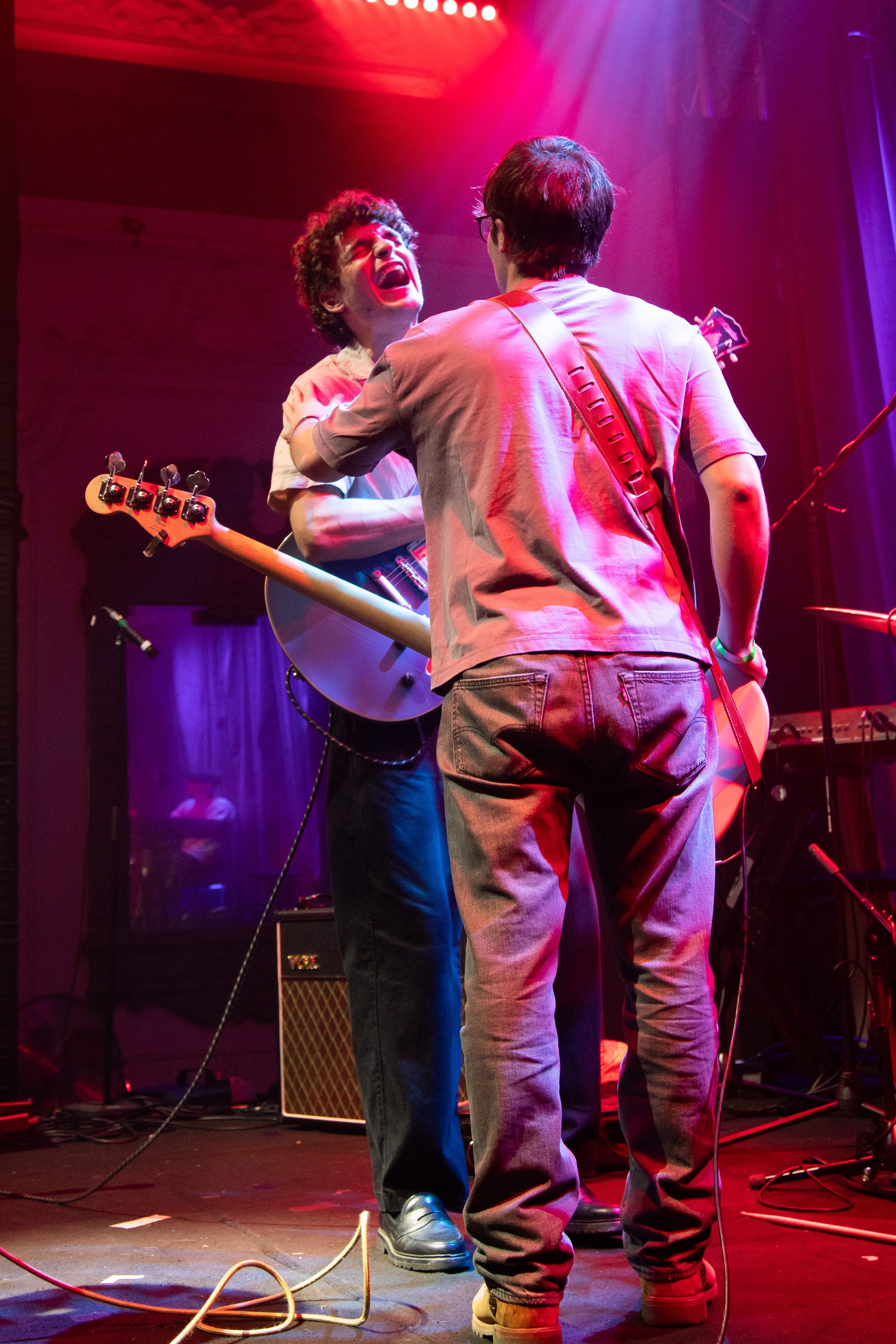 Two musicians on stage, one with curly hair playing an electric guitar, the other with straight hair and glasses singing passionately, illuminated by stage lights in pink and purple hues, with musical equipment and cables around them.
