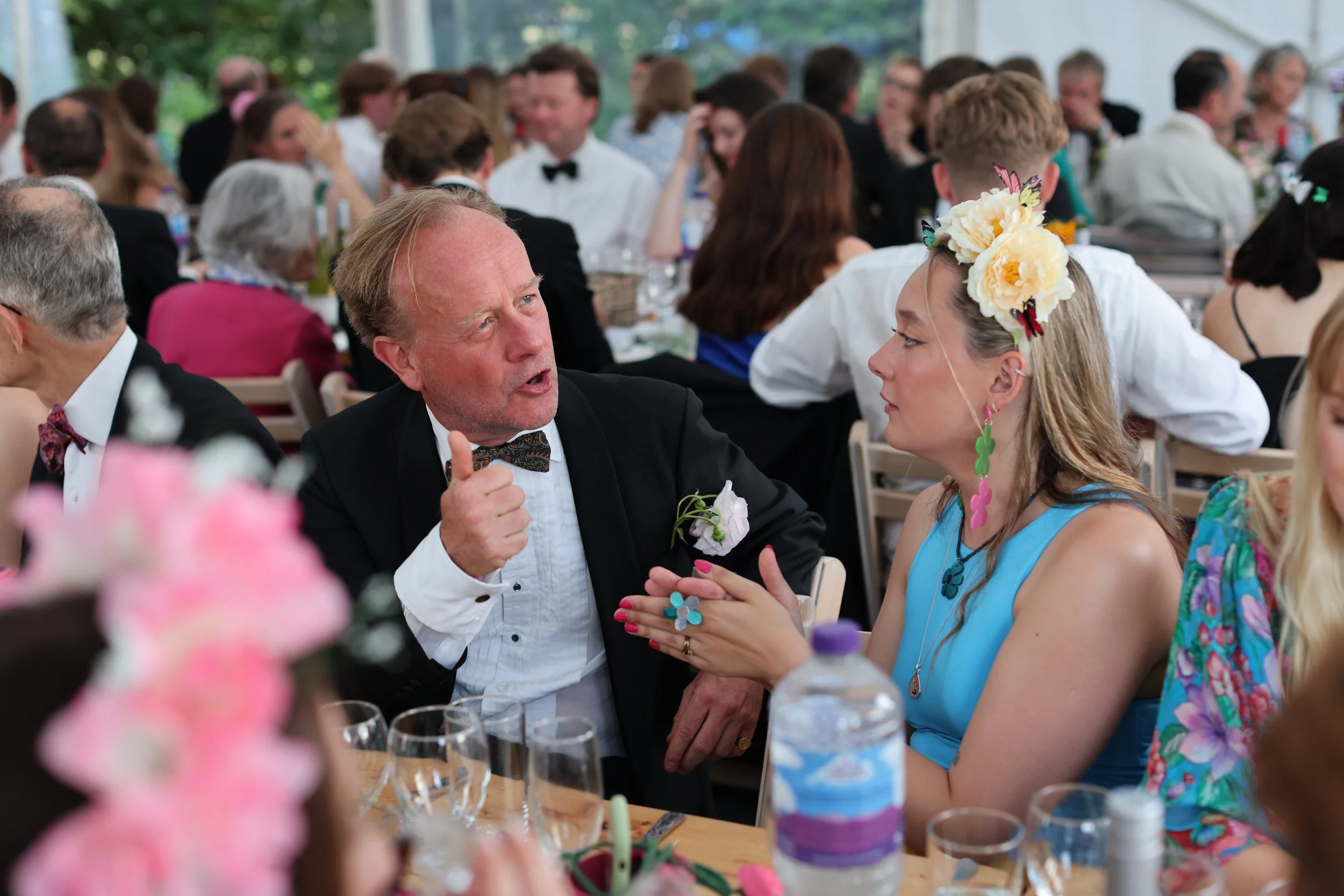 A group of people gathered at a lively social event, with a man and a woman engaged in conversation in the foreground. The woman is wearing a floral headpiece and colorful jewelry, and the man is in a tuxedo with a bowtie. The background shows other 