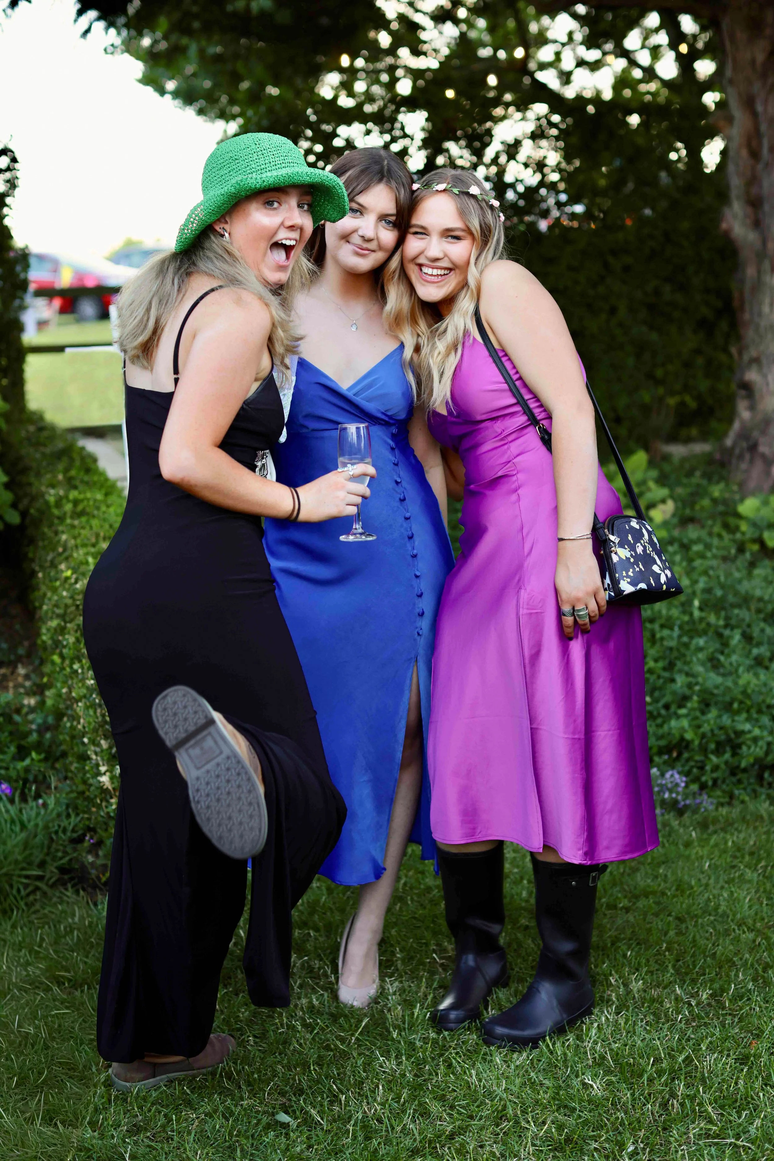 Three young women standing close together outdoors, smiling and posing for the camera in front of green trees, with blurry cars in the background. One wears a green hat, another a blue dress, and the third a purple dress with a floral headband.