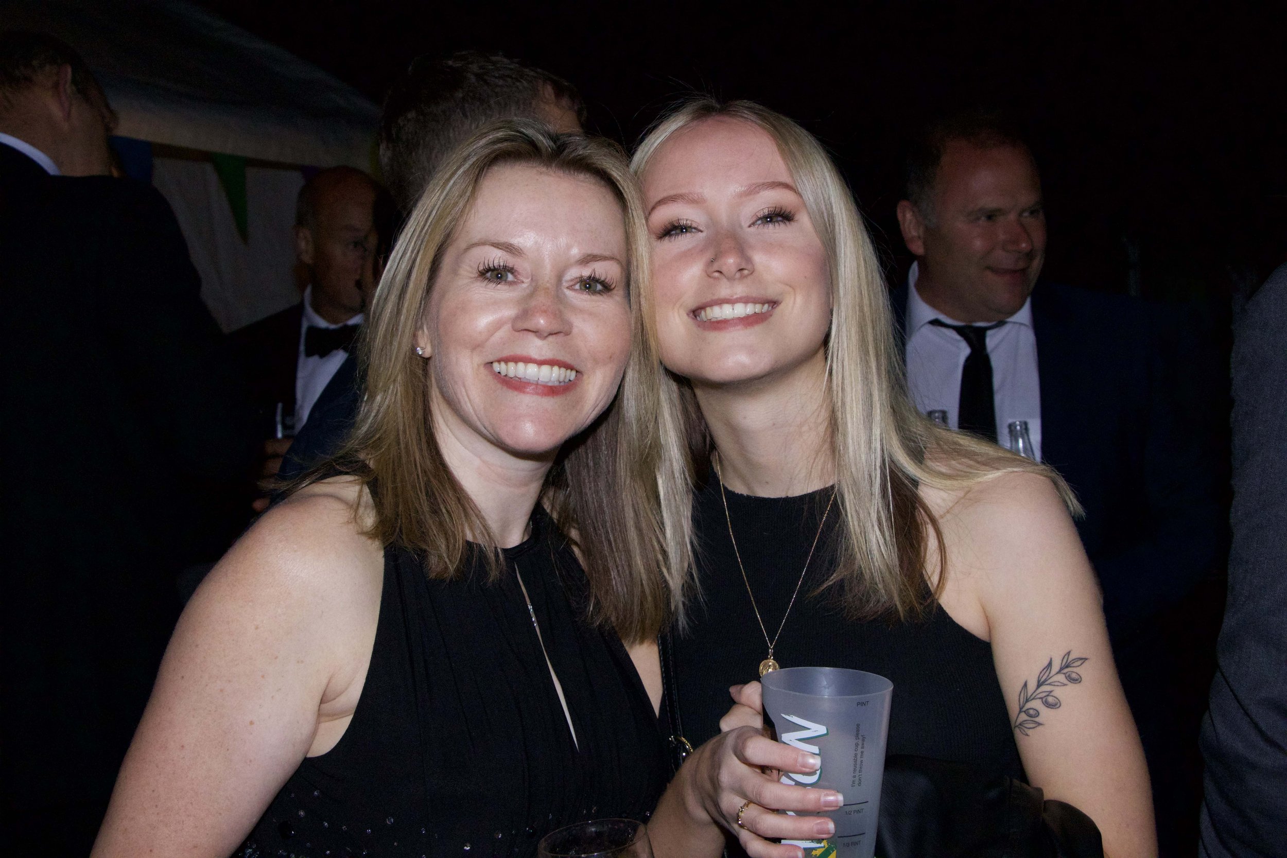 Two women smiling at a party, dressed in black, holding drinks, with other guests in formal attire in the background.