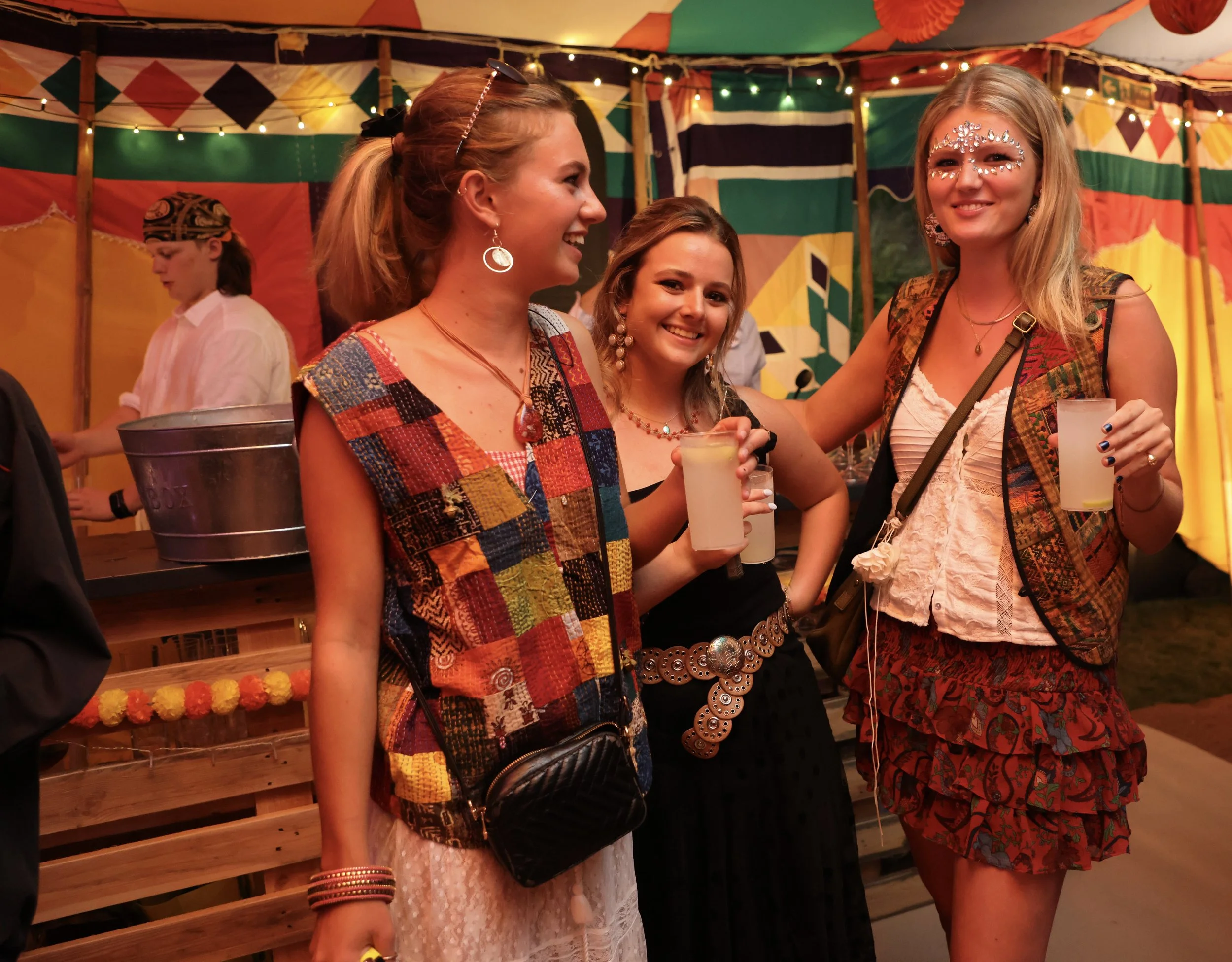 A group of women at an event or festival under a colorful canopy, smiling and holding drinks, with one woman wearing face jewels and colorful clothing.
