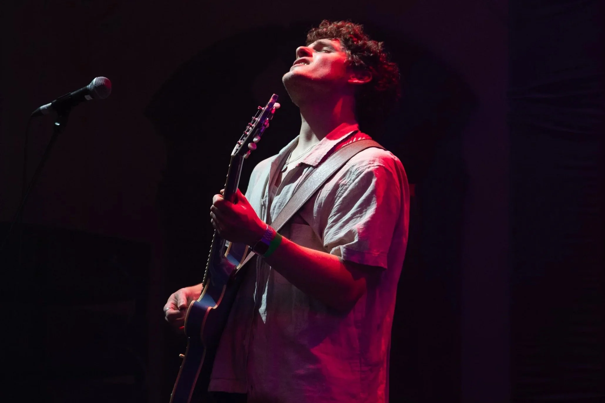 A young man with curly hair playing an electric guitar on stage with a dark background and red stage lighting.