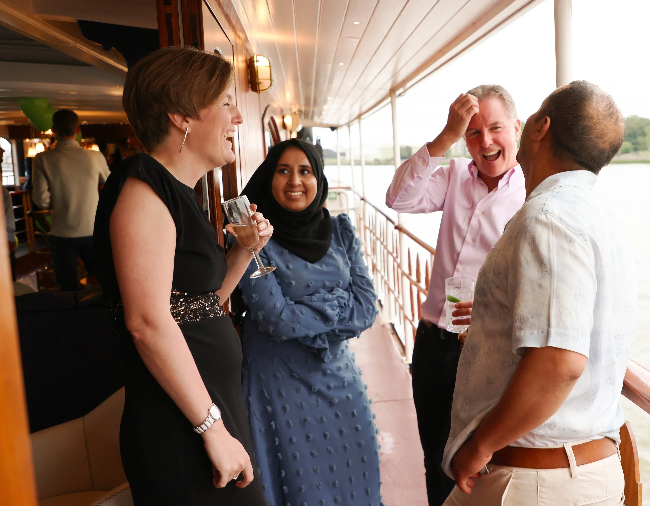 Four people are on a boat, smiling and talking. Two women and two men are visible, with one woman holding a glass of wine. They are dressed casually, and the background shows water and sky.